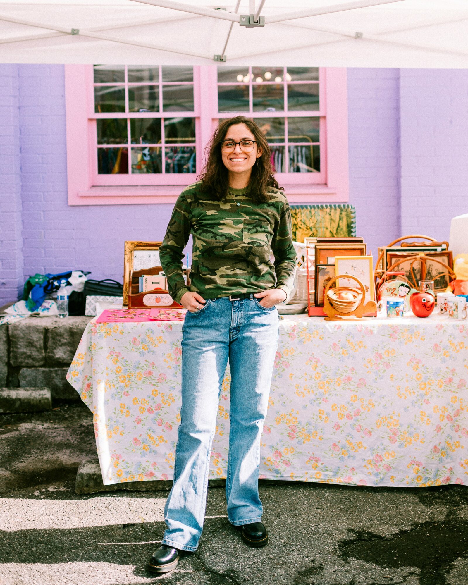 Woman with glasses and long hair stands in front of a table with colorful items, outdoors with a purple and pink building behind.