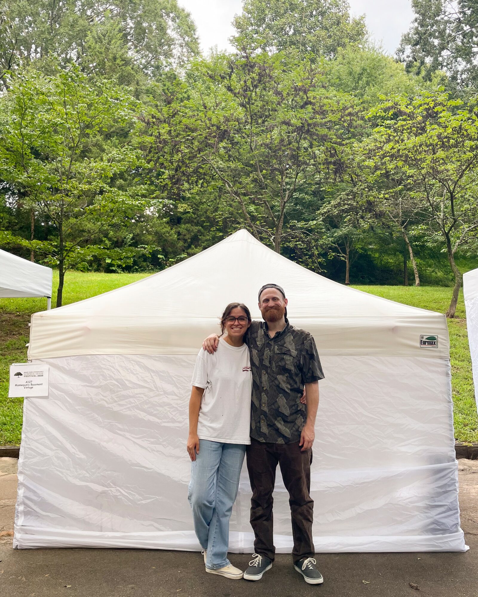 Two people stand in front of a white tent outdoors, surrounded by green trees and grass.