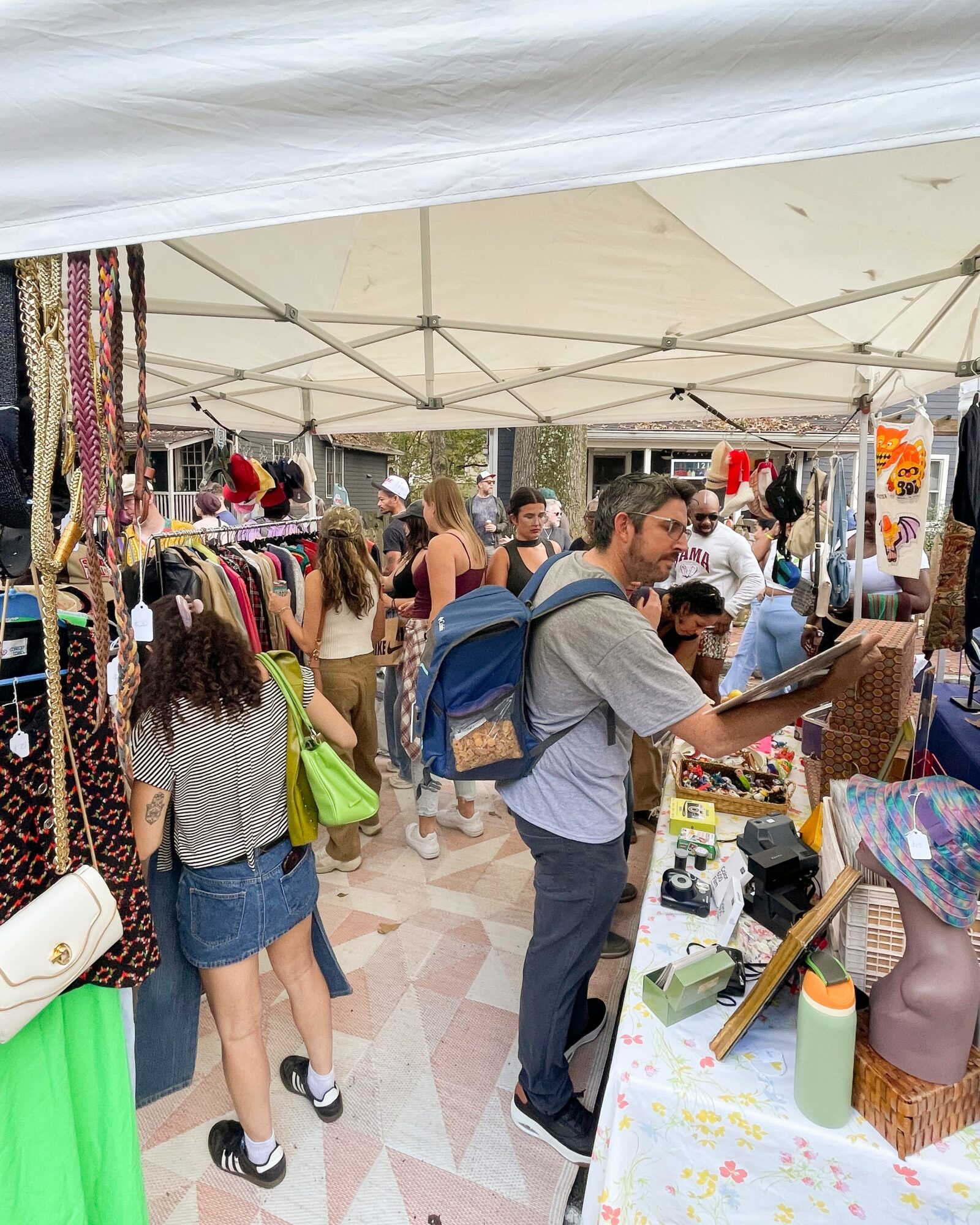 People browsing outdoor market stalls under a white canopy, with clothing and accessories on display, and shoppers interacting.