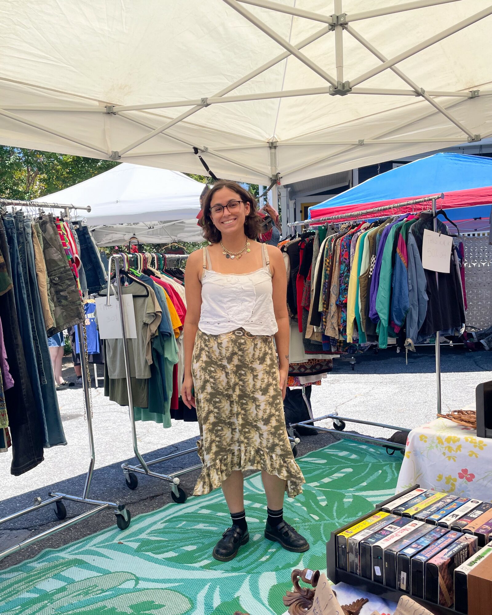 Woman standing under a tent at a market, surrounded by clothing racks and a table with books, smiling.
