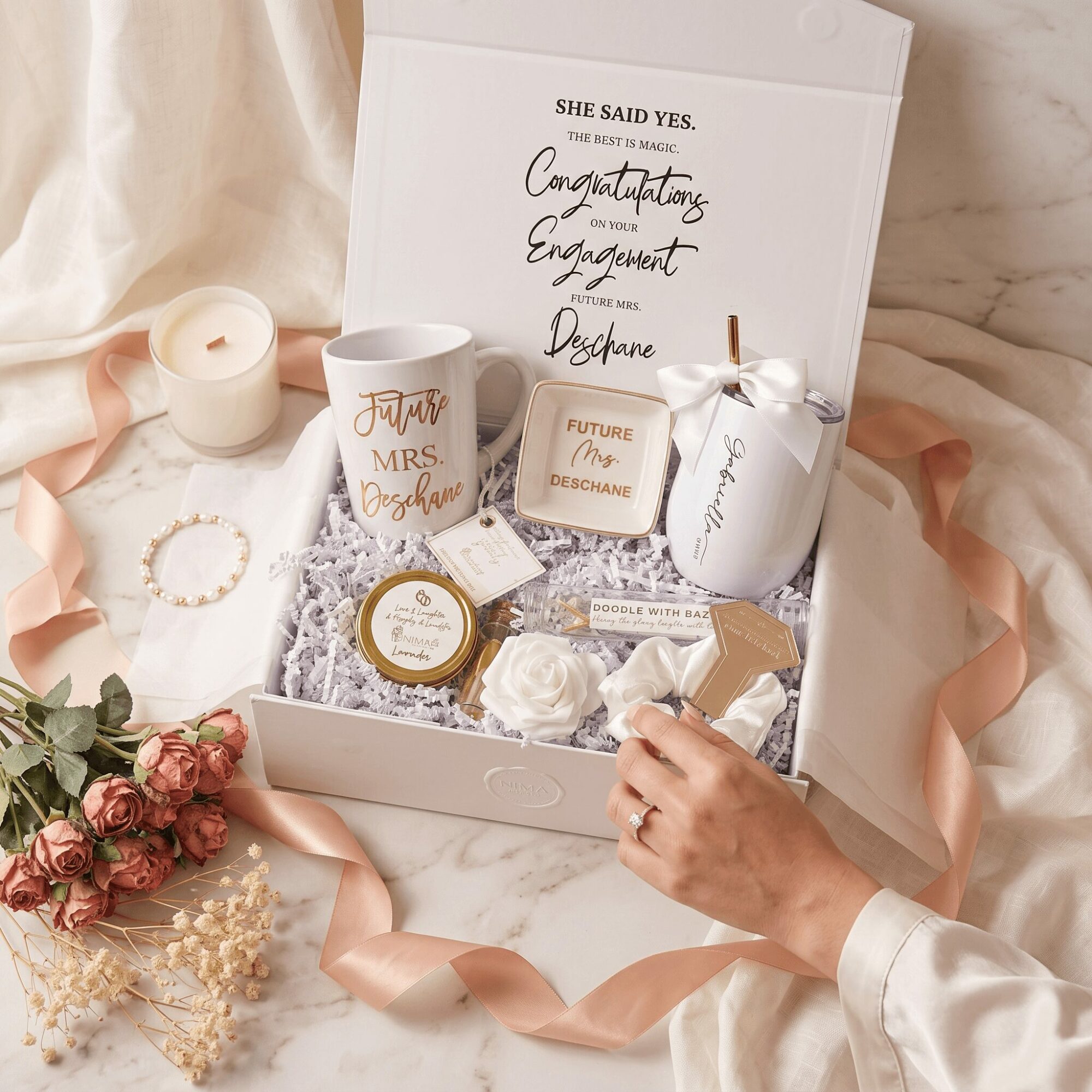 Open gift box with wedding items, surrounded by flowers and ribbons, on a soft fabric surface.
