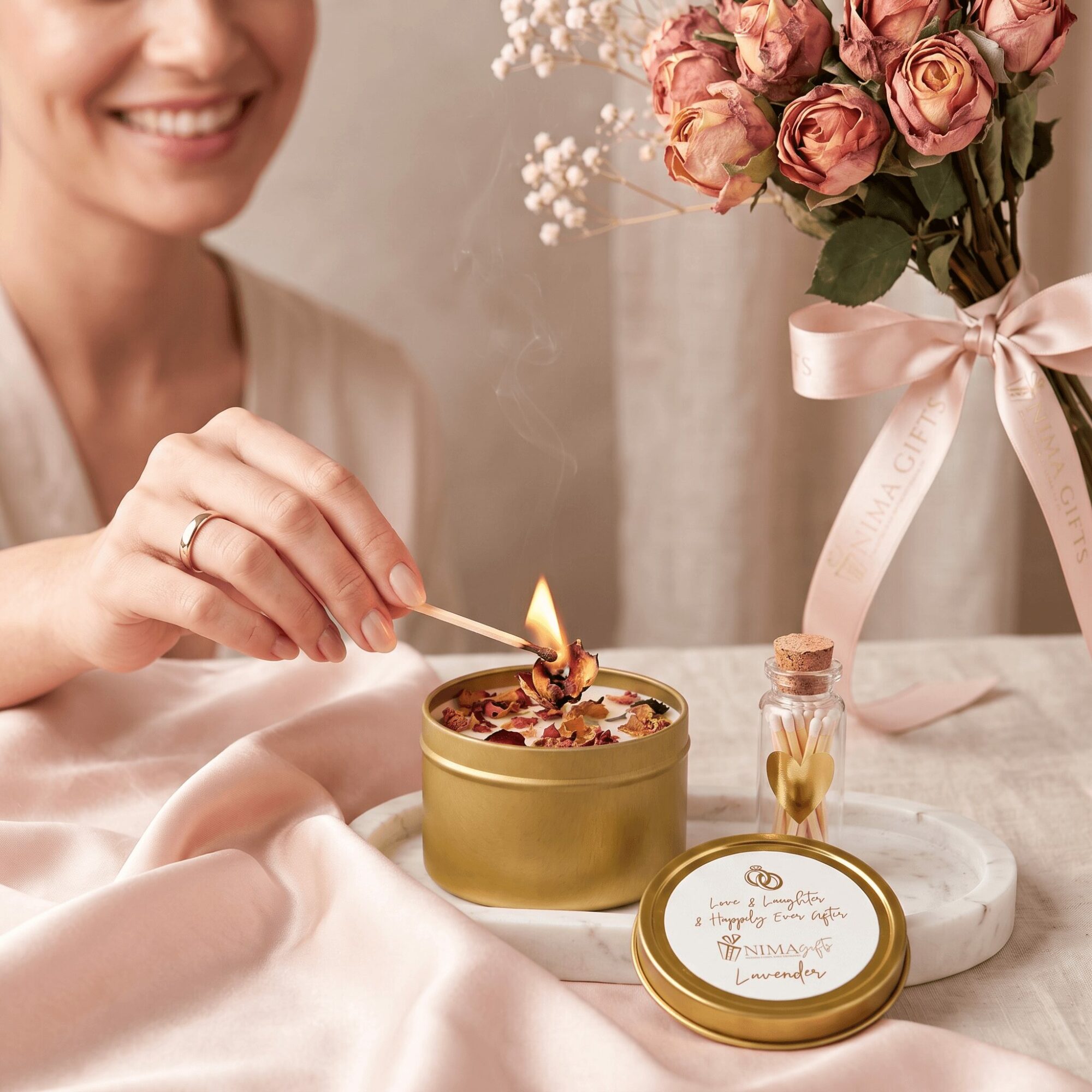 Person lighting a candle with a match, smiling, with a bouquet of pink roses in a vase nearby, on a table.