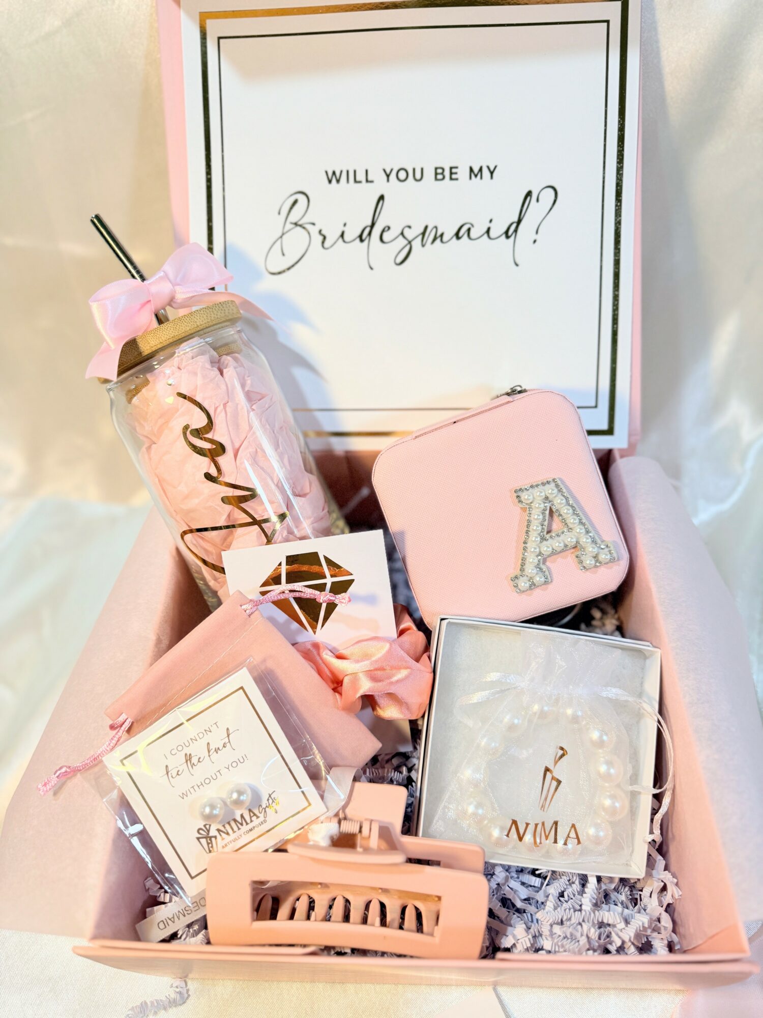 Pink gift box with various items including a water bottle, a small purse with letter A, and a card, with a sign asking, 'Will you be my Bridesmaid?'.