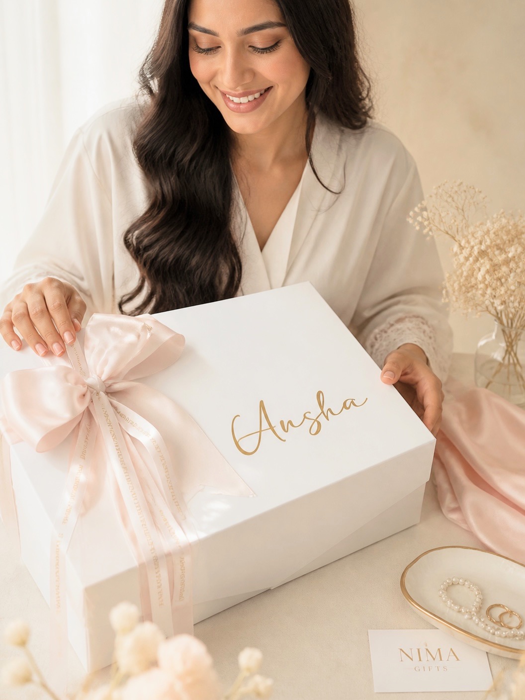 Smiling woman opening a gift box with a pink bow, with the name 'Ansha' written on it, in a cozy setting.