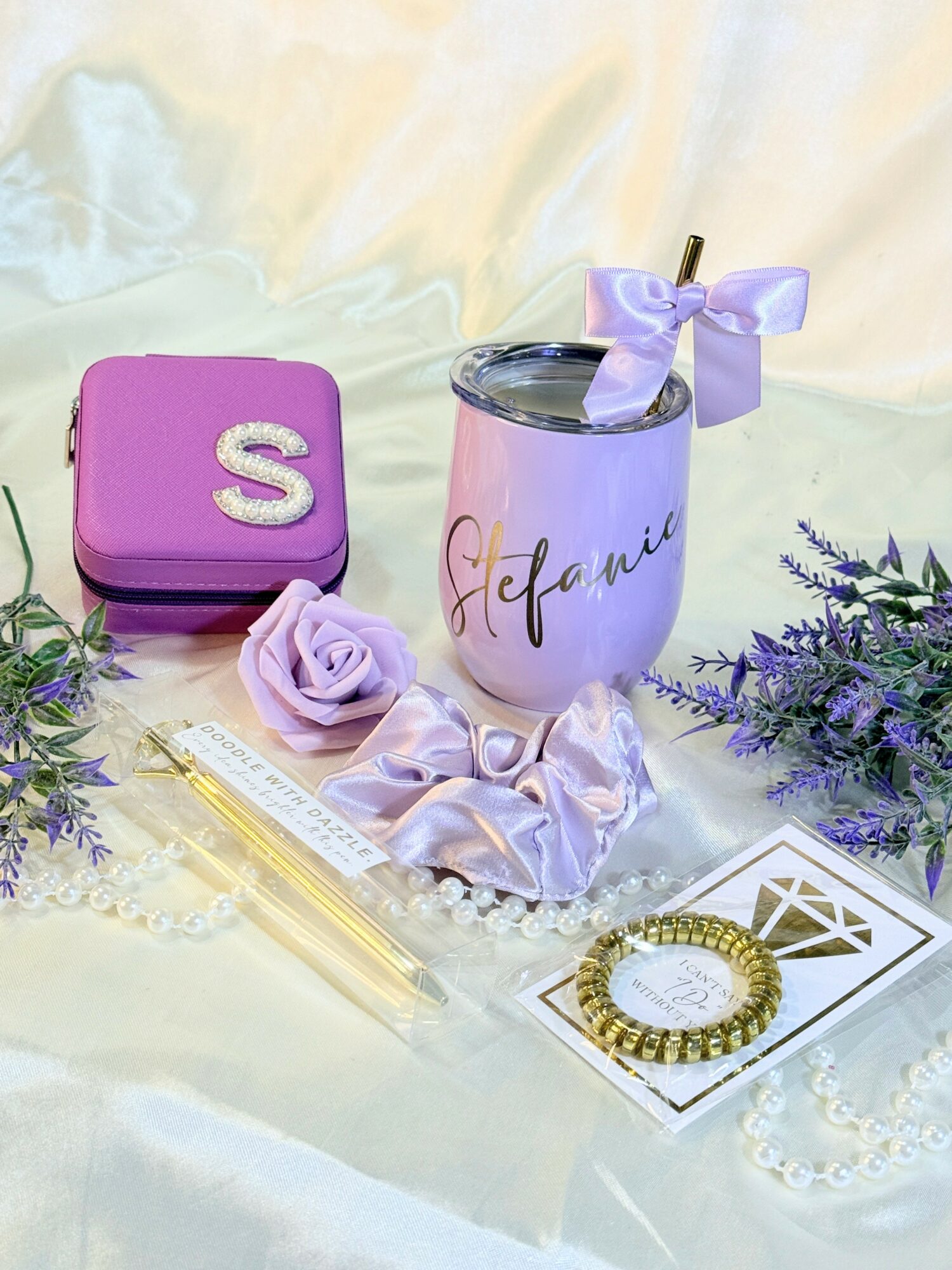 Pink tumbler with purple bow, purple purse with letter 'S', purple flower, gold bracelet, and purple flowers on satin fabric.