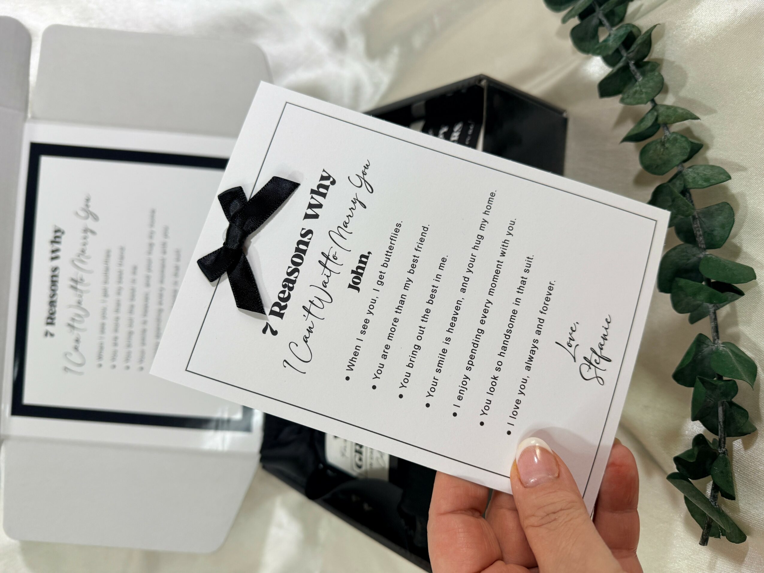 Person holding a card with a black ribbon, surrounded by eucalyptus leaves, with a box and framed document in the background.