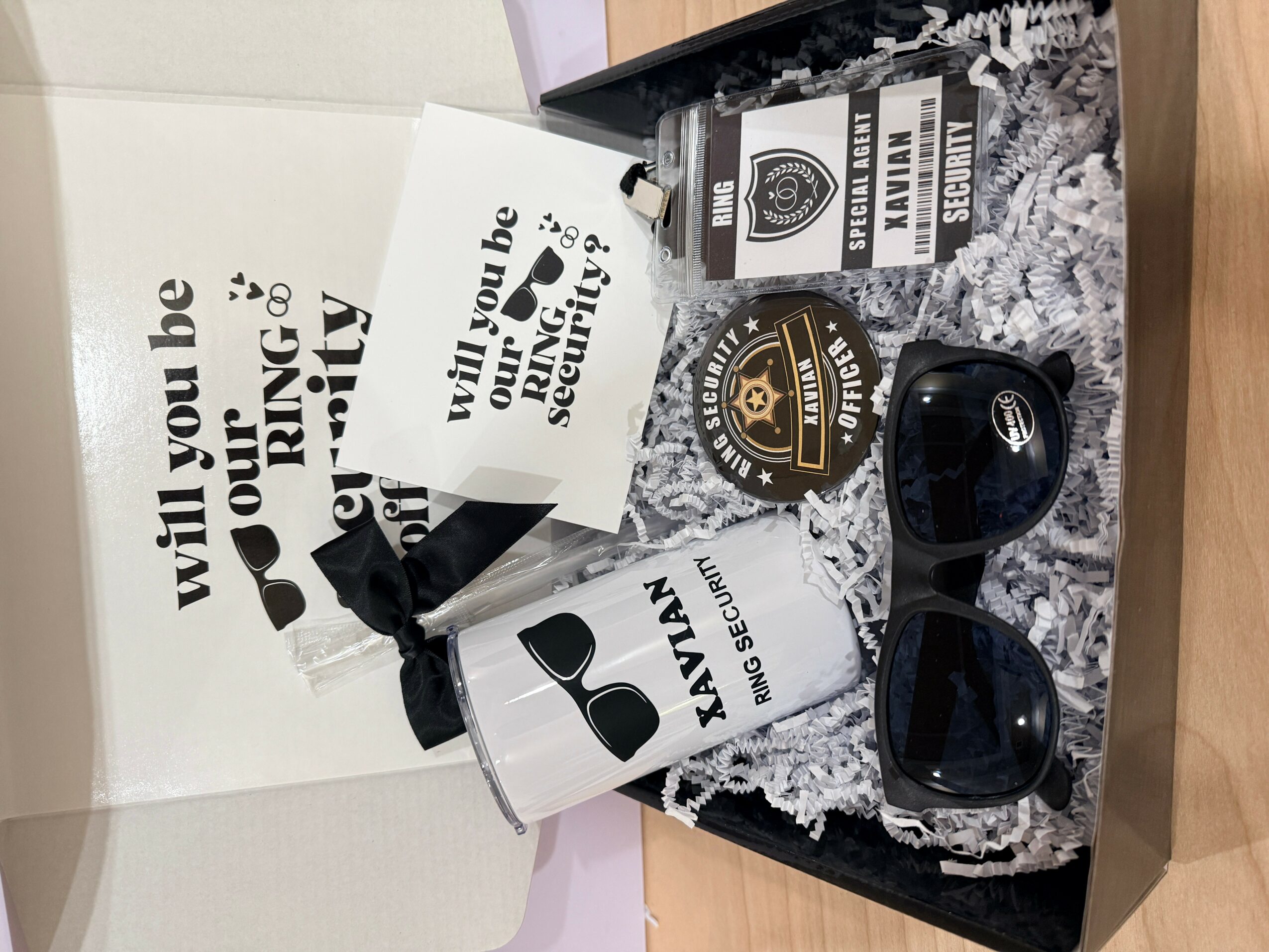 Open box with sunglasses, a small bottle, and printed materials on a wooden surface.