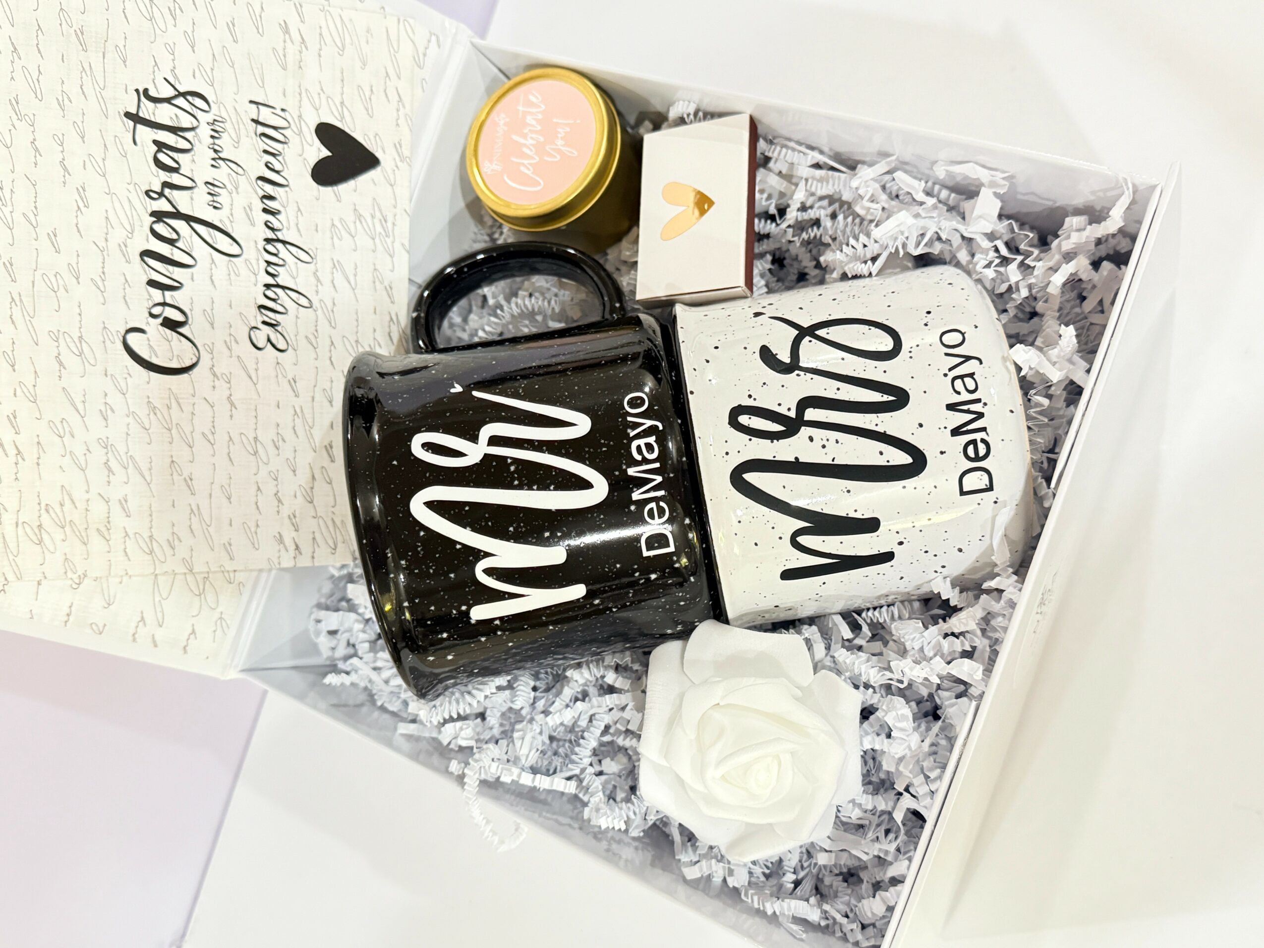 Gift box with black mug, white mug, white rose, small jar, and card, arranged on shredded paper, celebrating a special occasion.