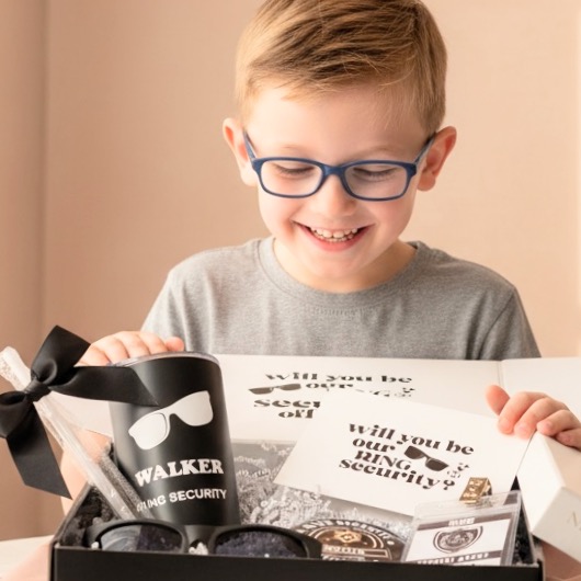 Child with glasses smiling at a security-themed gift box, surrounded by security-related items, indoors with beige background.
