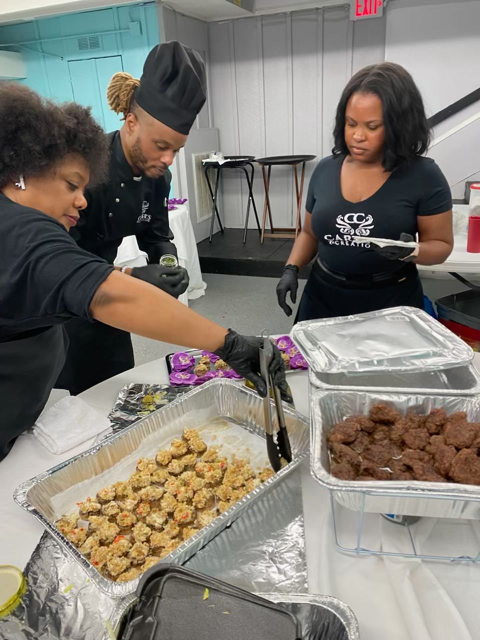Two women preparing food at a table with trays of cooked items, one woman wearing black gloves, in a kitchen setting.