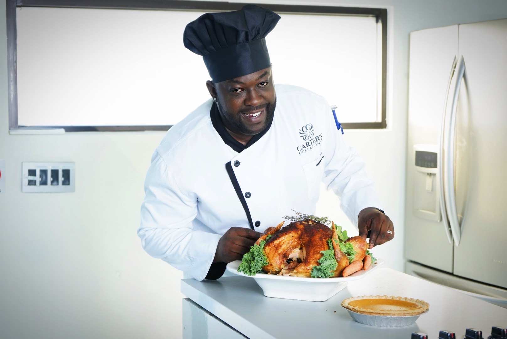 Chef in uniform and hat holding a roasted turkey on a plate in a kitchen, smiling at the camera.