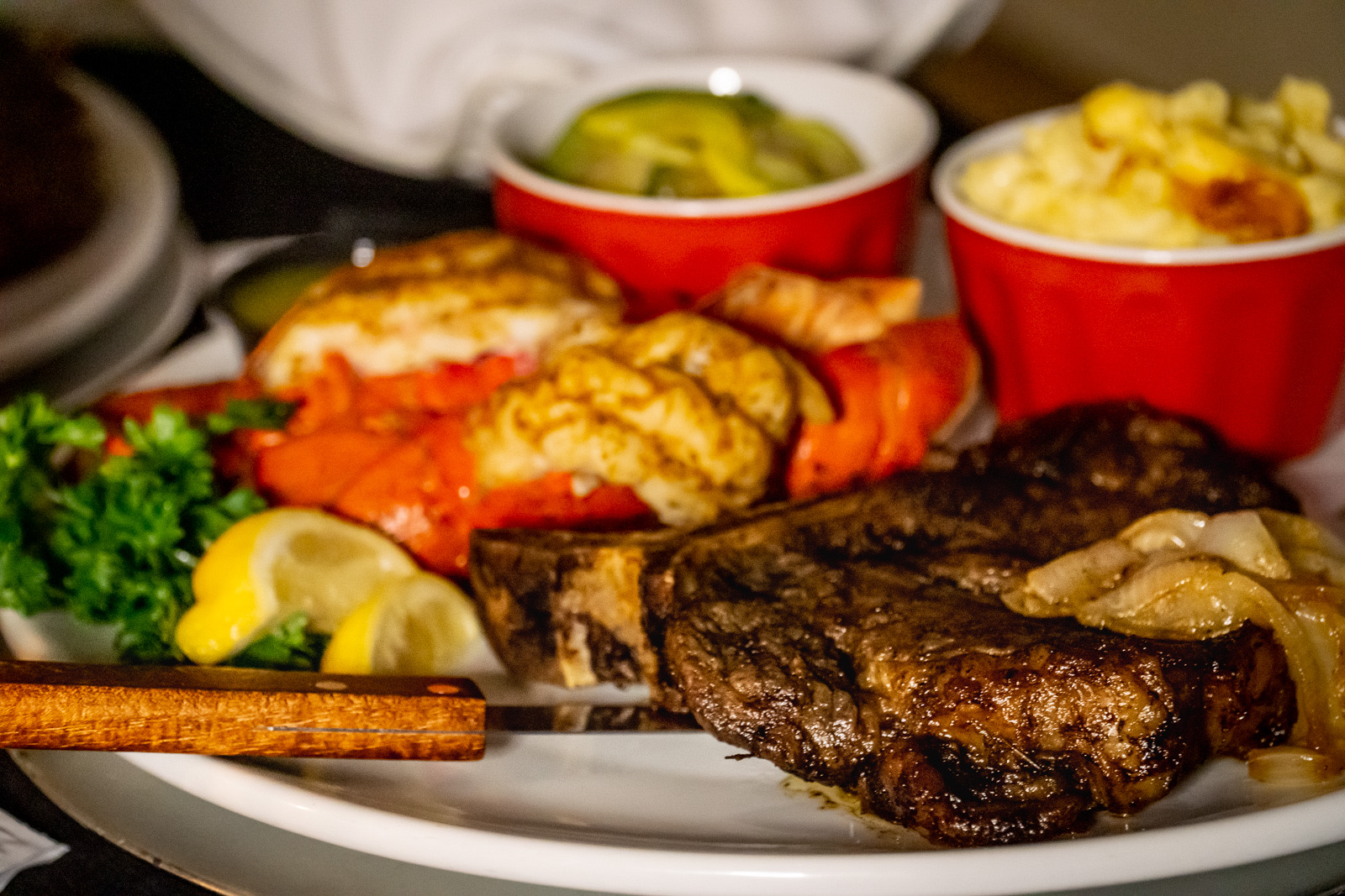 Plate with grilled steak, fried chicken, vegetables, and two bowls of side dishes, garnished with lemon and parsley.
