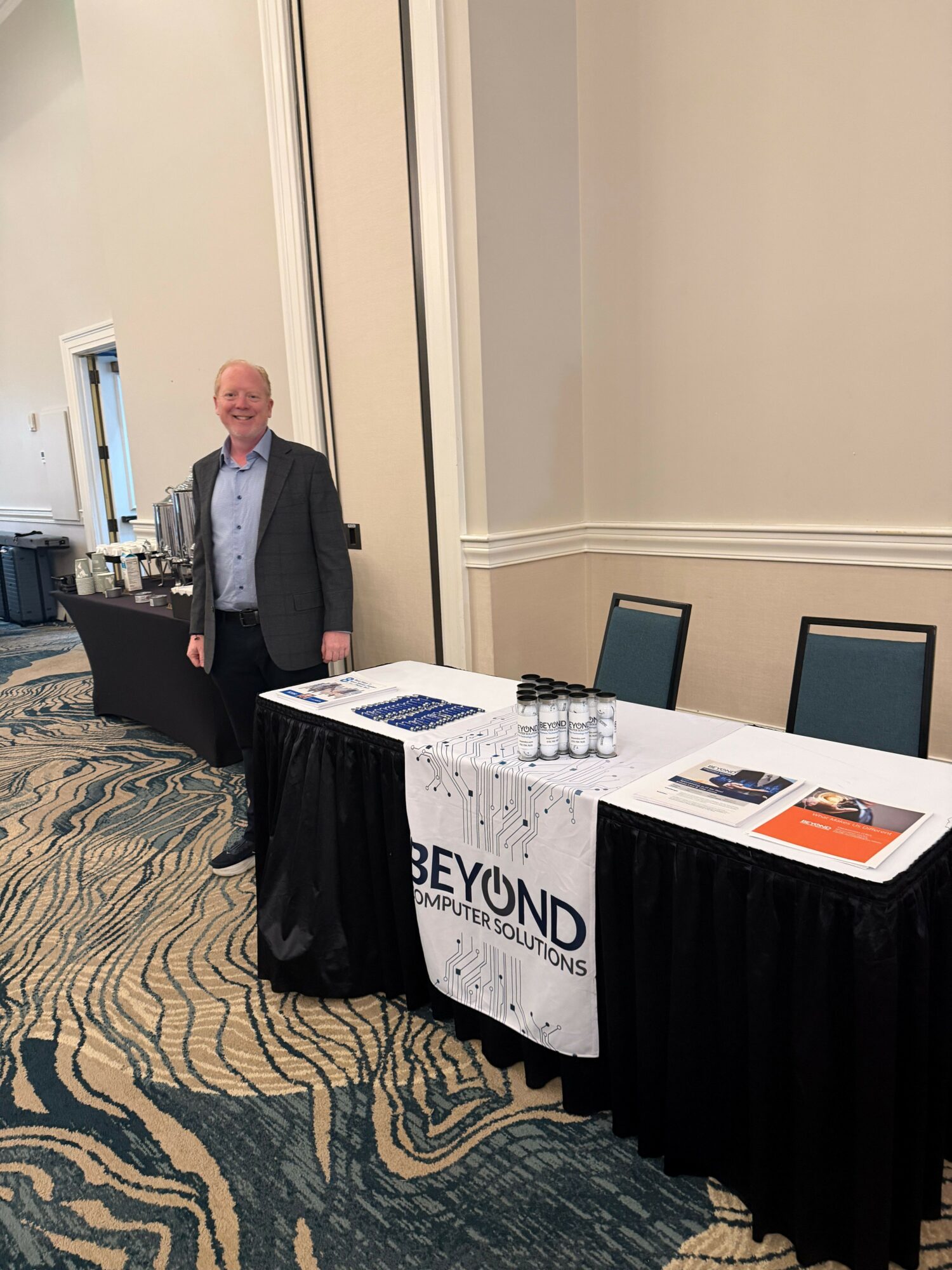 Man standing behind a table with promotional materials in a conference room, carpeted floor, and a window in the background.