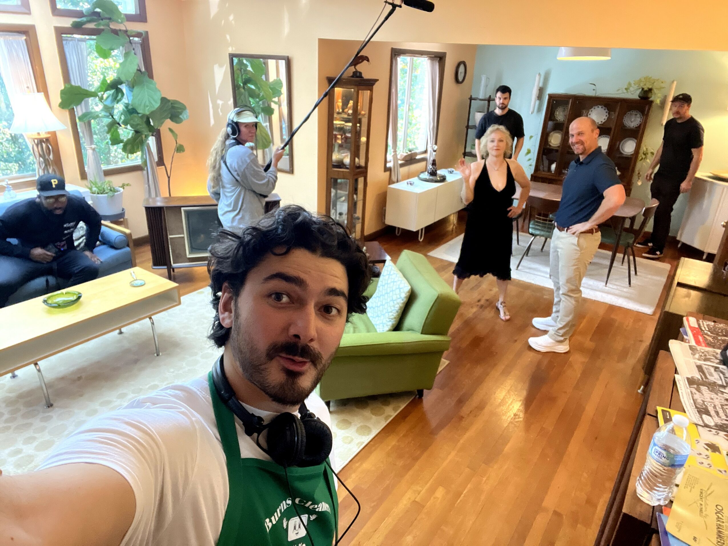 Group of people in a living room, some talking, one person filming, and others sitting or standing, with wooden floors and furniture.