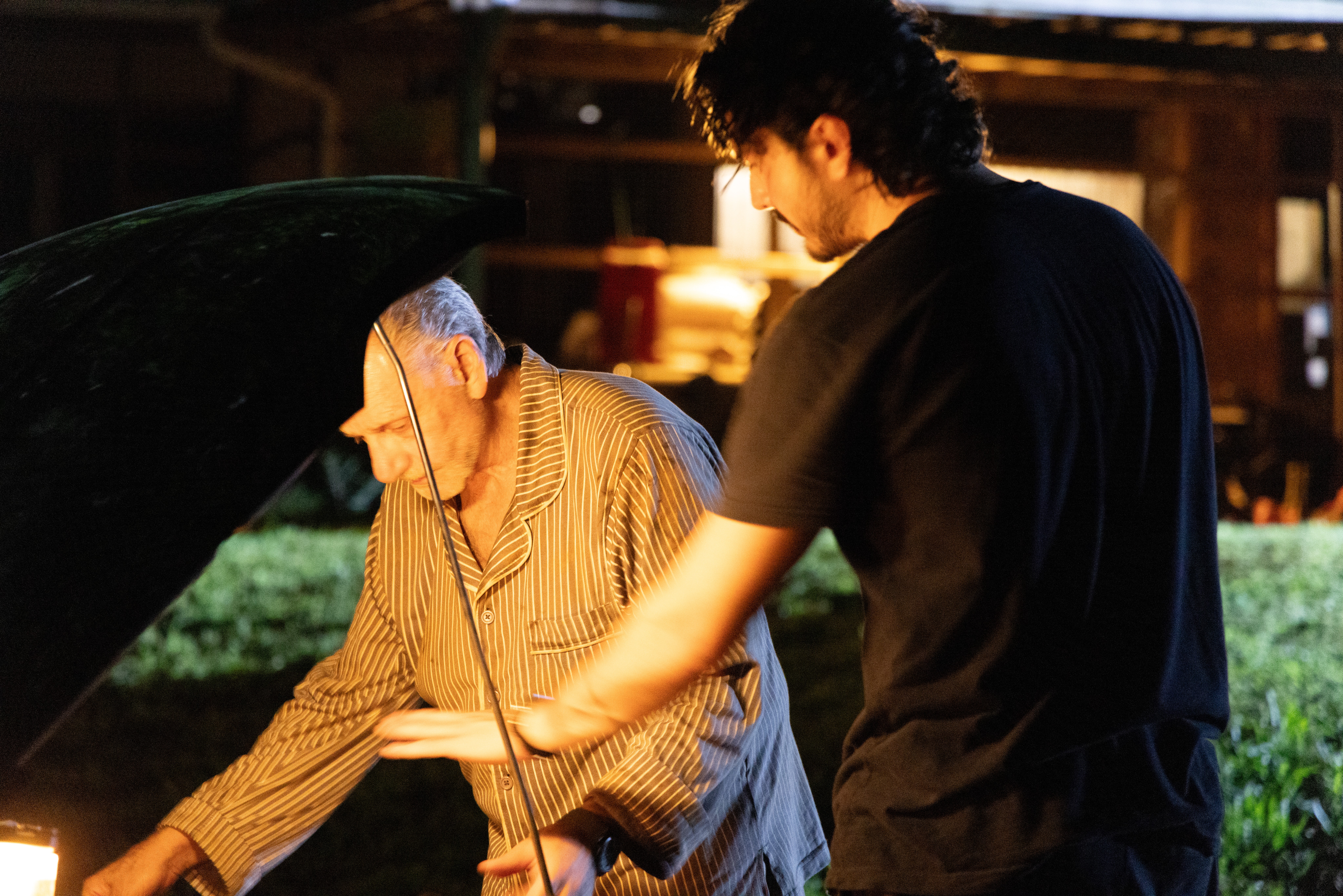 Two people examining a car engine outdoors at night, one older with gray hair, the other younger with dark hair.