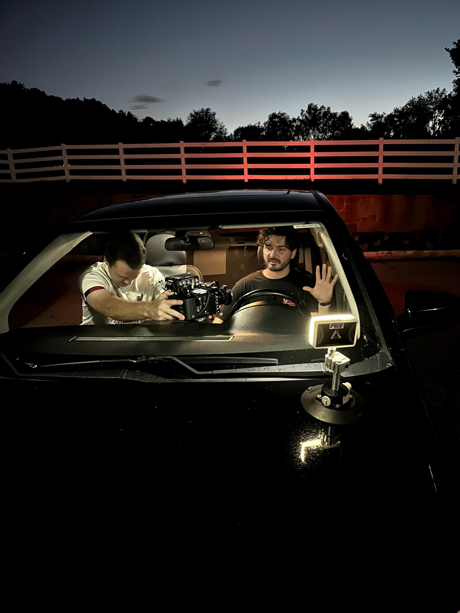 Two people inside a car at night, one is filming with a camera, the other is waving, with a bridge and trees outside.