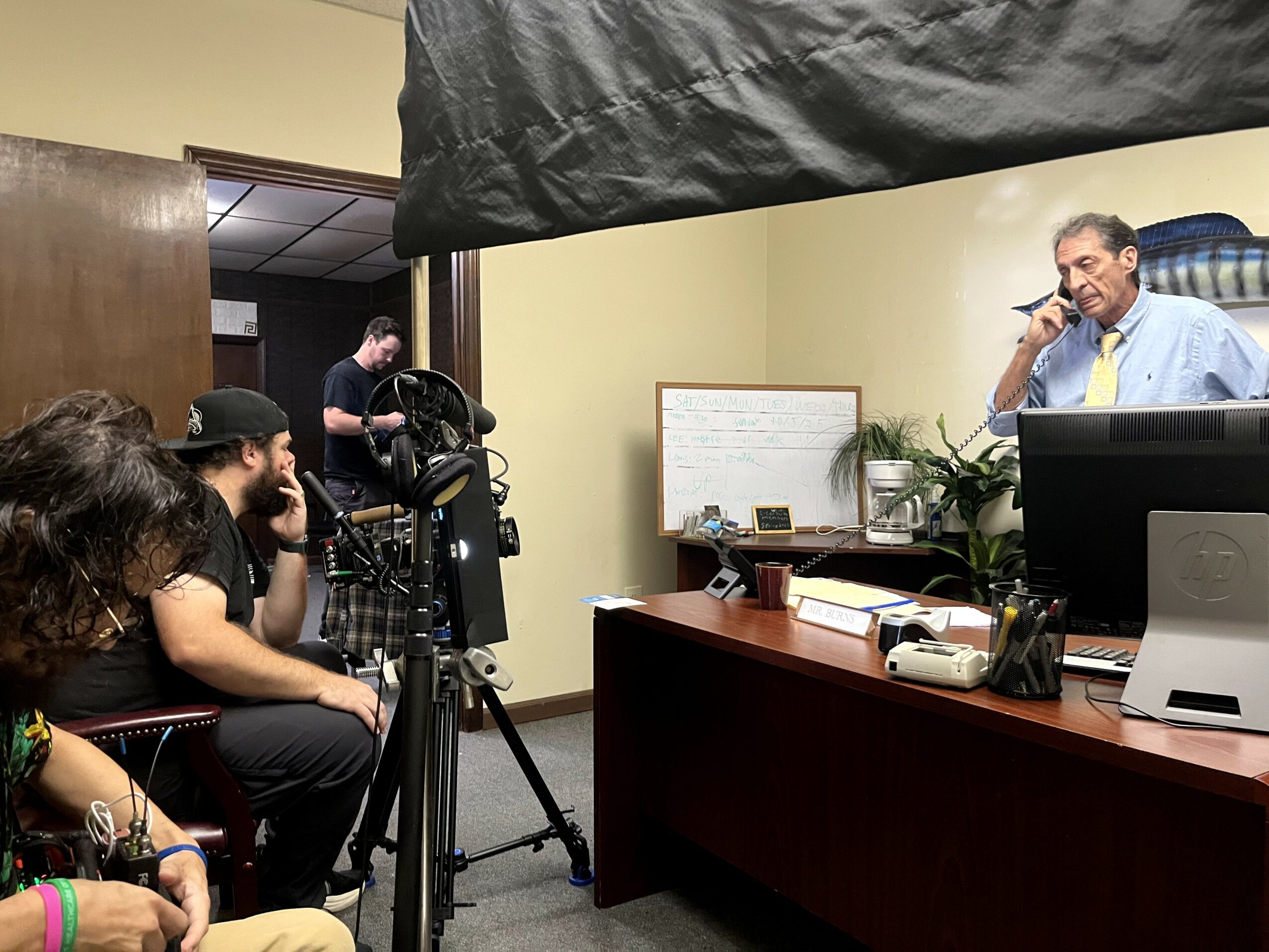 People seated in a room watching a man at a desk with a whiteboard and computer, filming equipment nearby.
