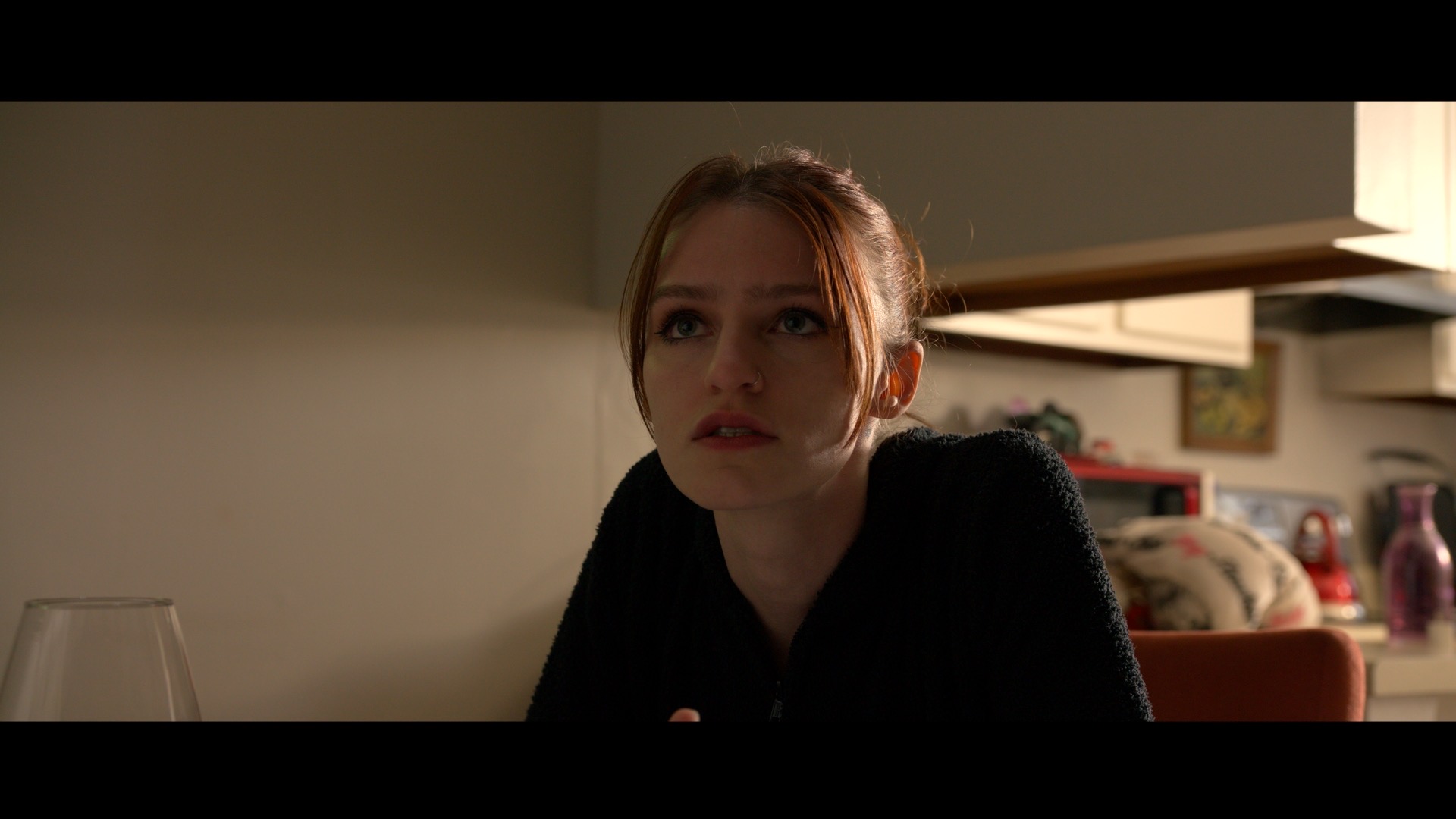 Young woman with light brown hair and black top looks upward in a dimly lit room.