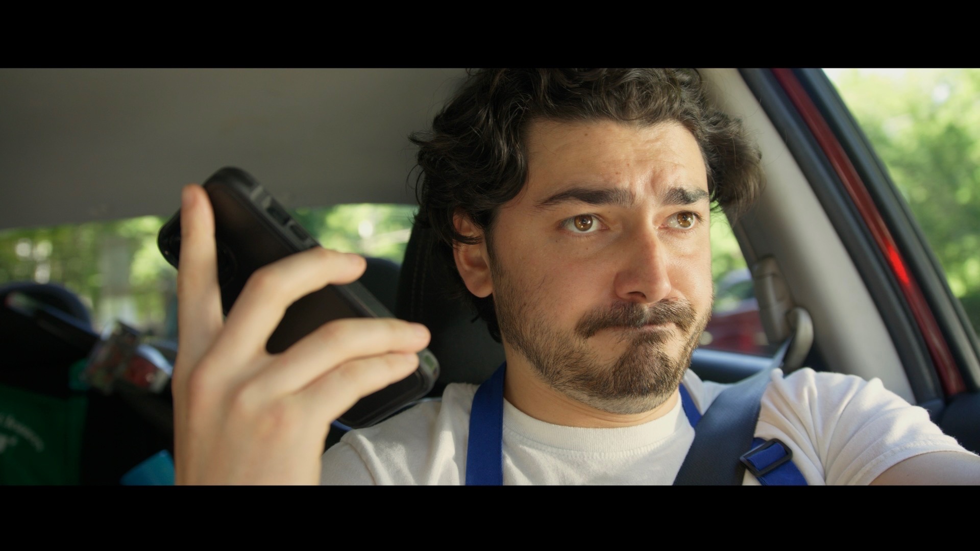 Man with curly hair and beard looking at phone while driving a car, with a serious expression.