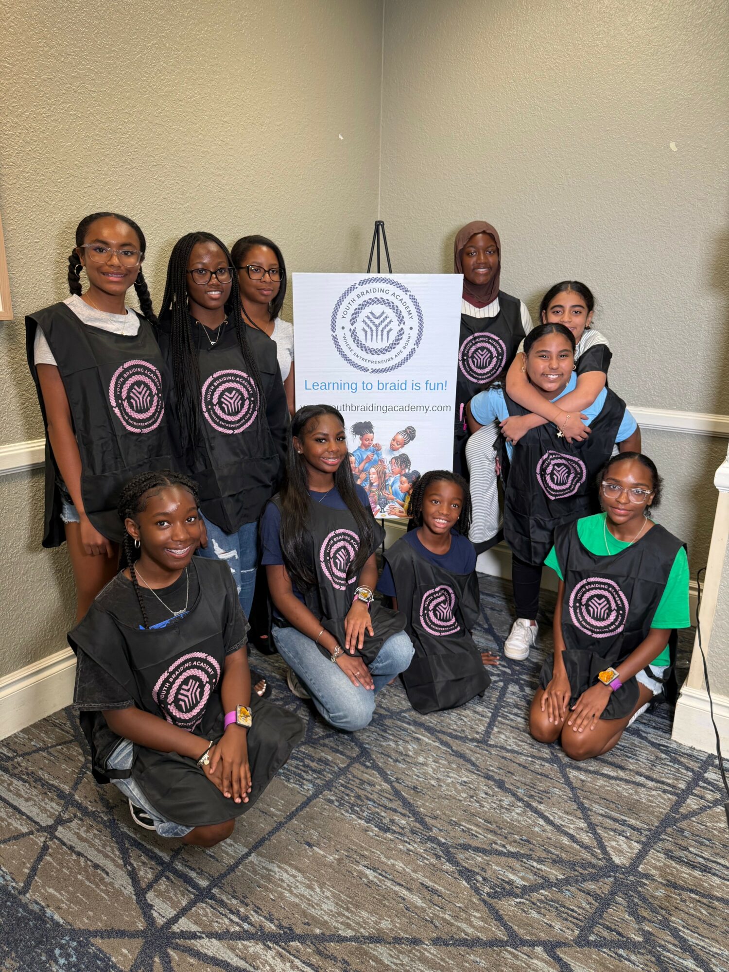 Group of ten children and teenagers posing indoors with a banner behind them, some kneeling and some standing.