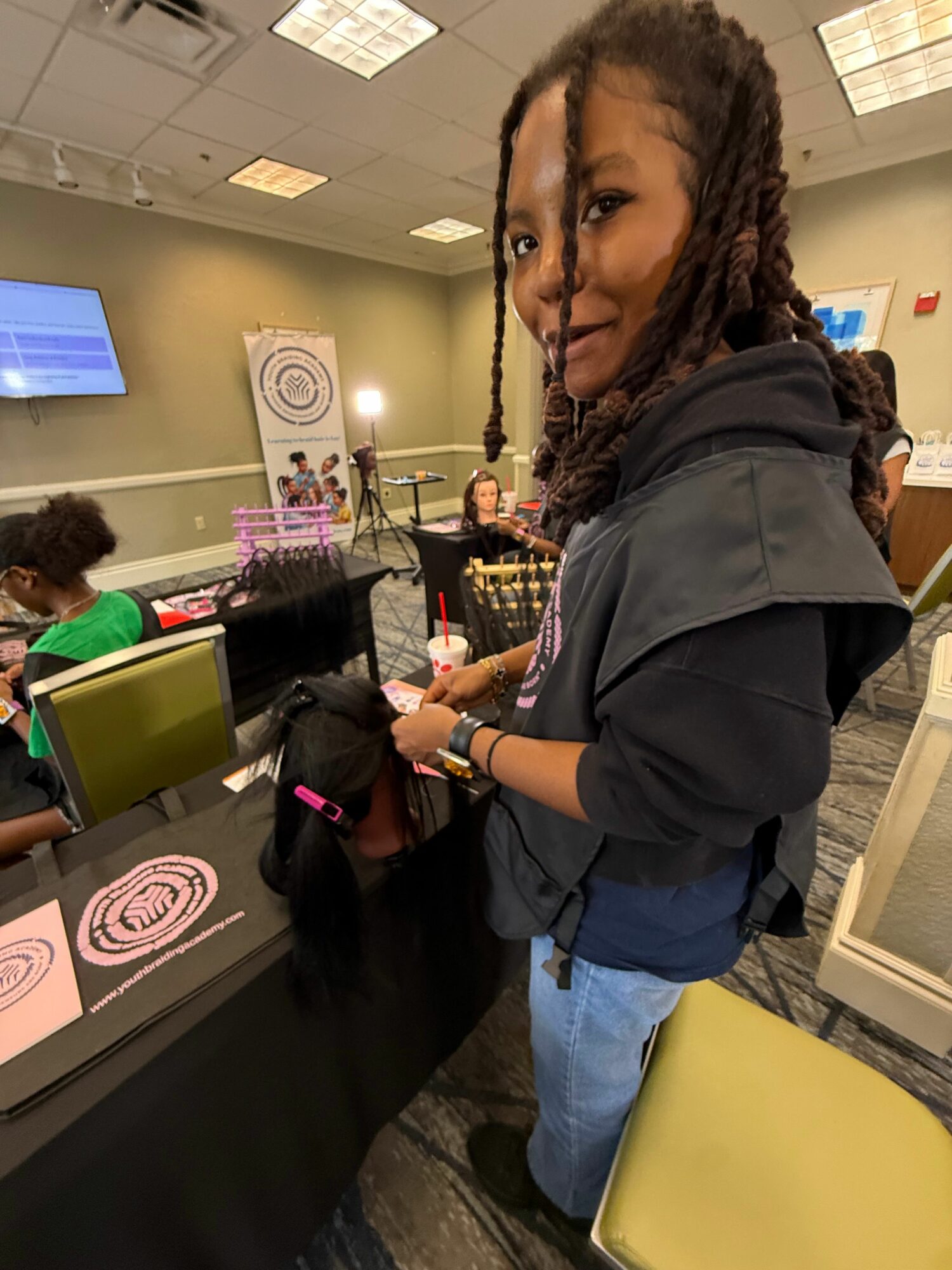 Woman with braided hair and glasses standing at a table with pink and black items, in a room with yellow chairs and a computer monitor.