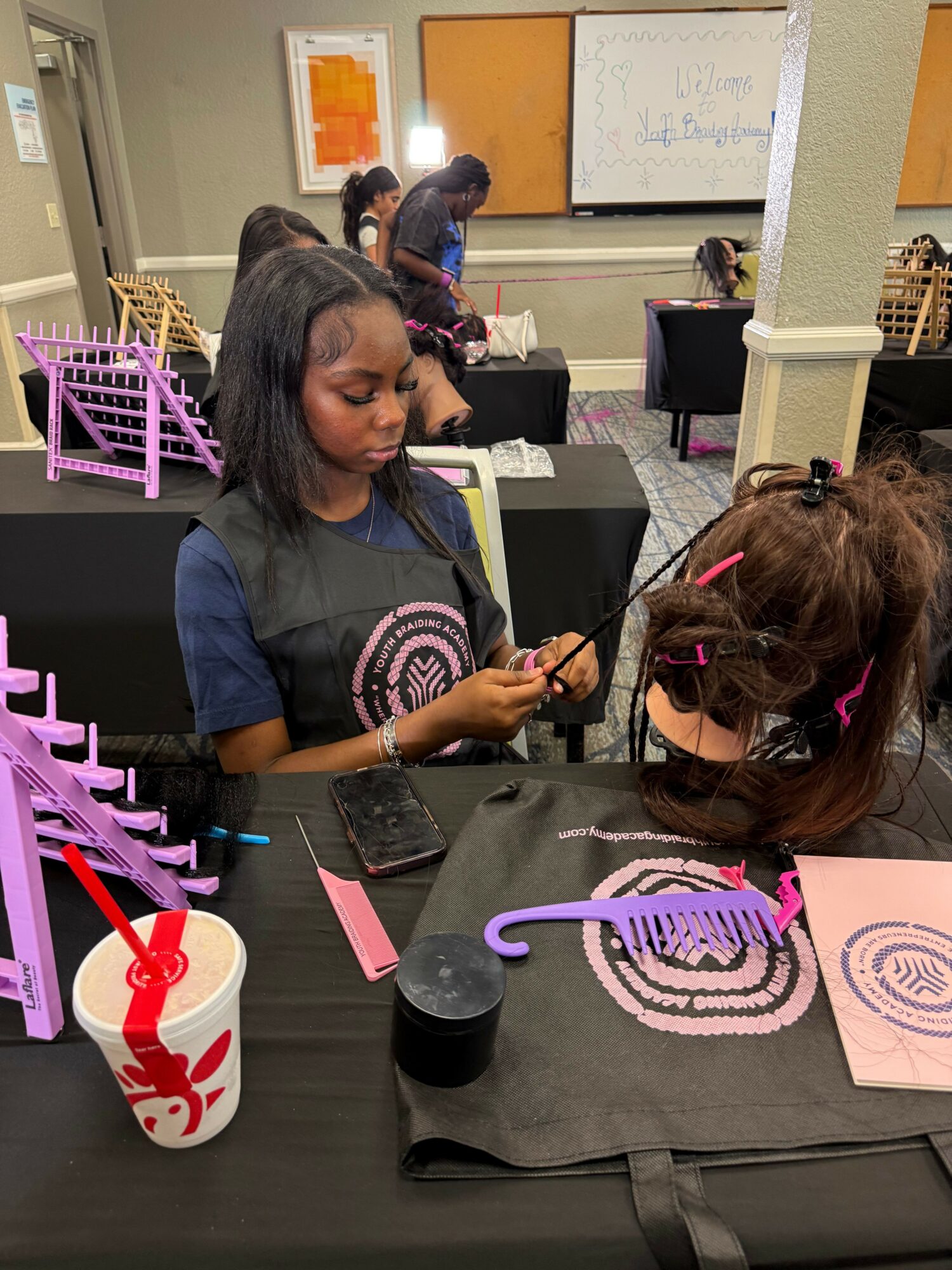 Two children with long hair, one with a pink hairband, sit at a table with a whiteboard and classroom supplies, engaging in activity.