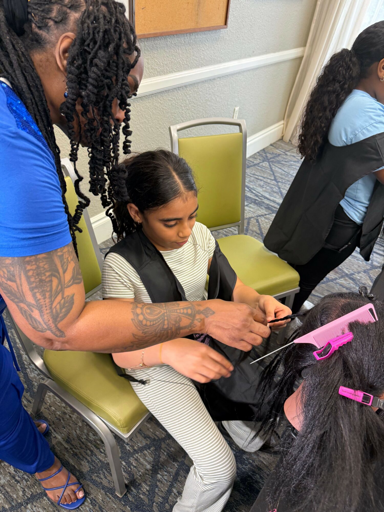 Person with tattoos on arm using a curling iron on long dark hair, seated at a table with pink hair tools nearby.