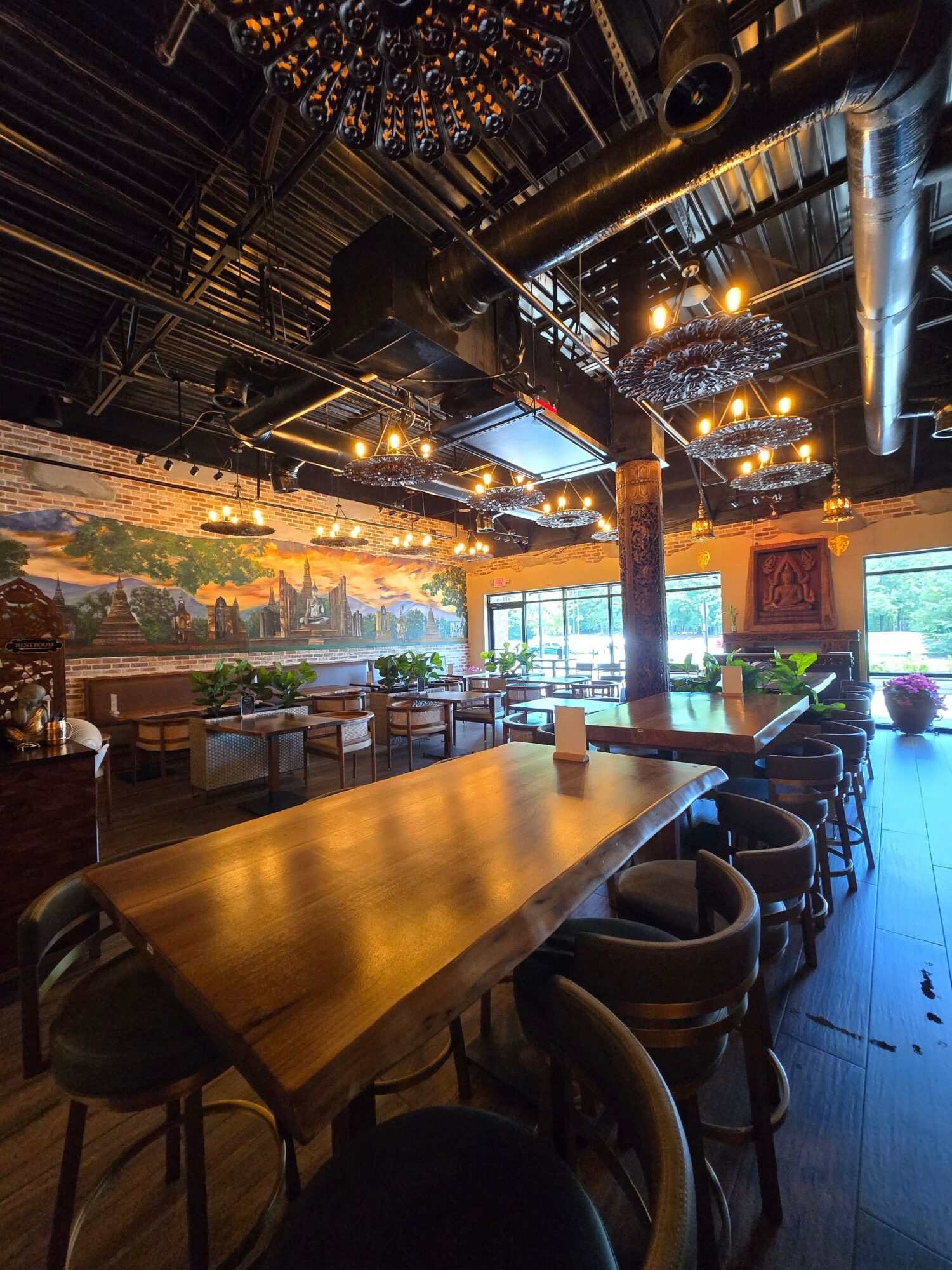 Interior of a restaurant with a long wooden table, chairs, large windows, and decorative lighting fixtures.