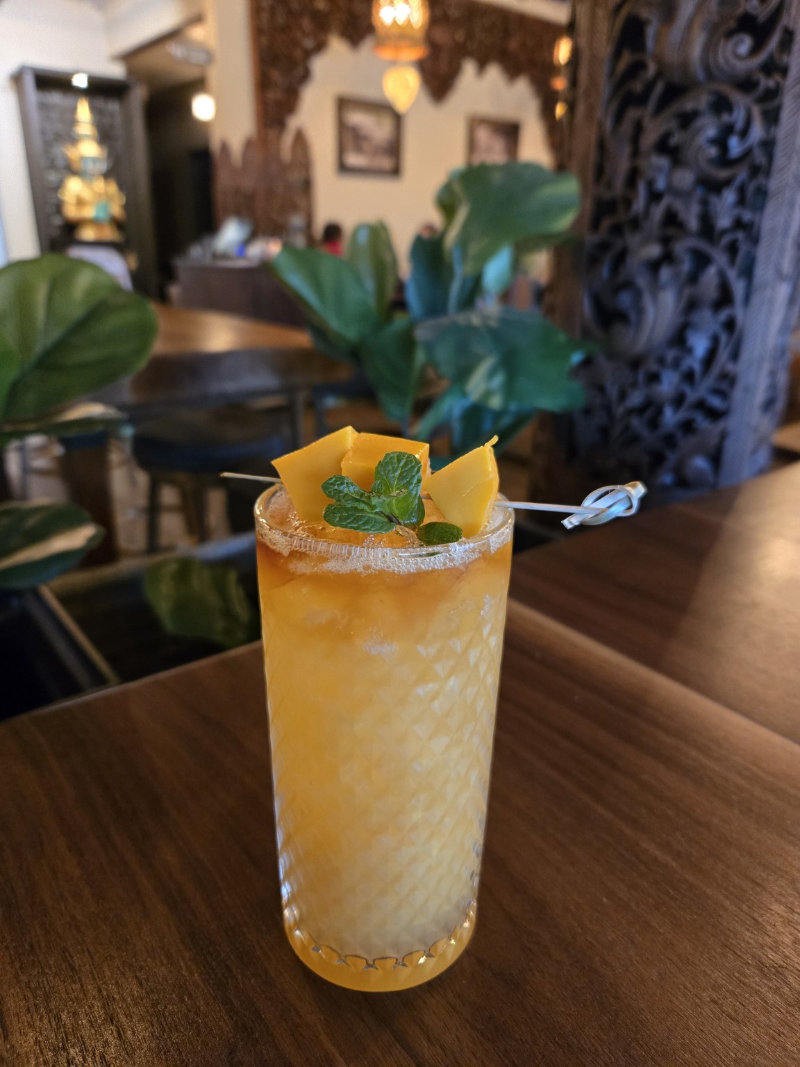 Tall glass of layered yellow and white drink garnished with mint and lemon peel, on a wooden table in a cozy restaurant.