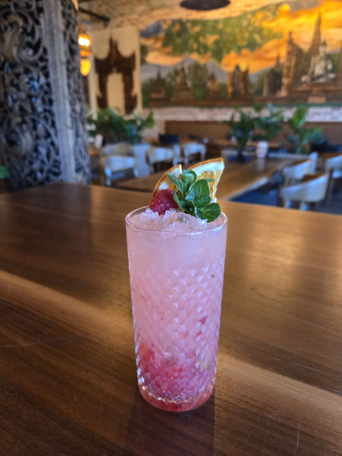 Pink cocktail garnished with mint, lemon slice, and strawberry on a wooden table in a decorated restaurant.