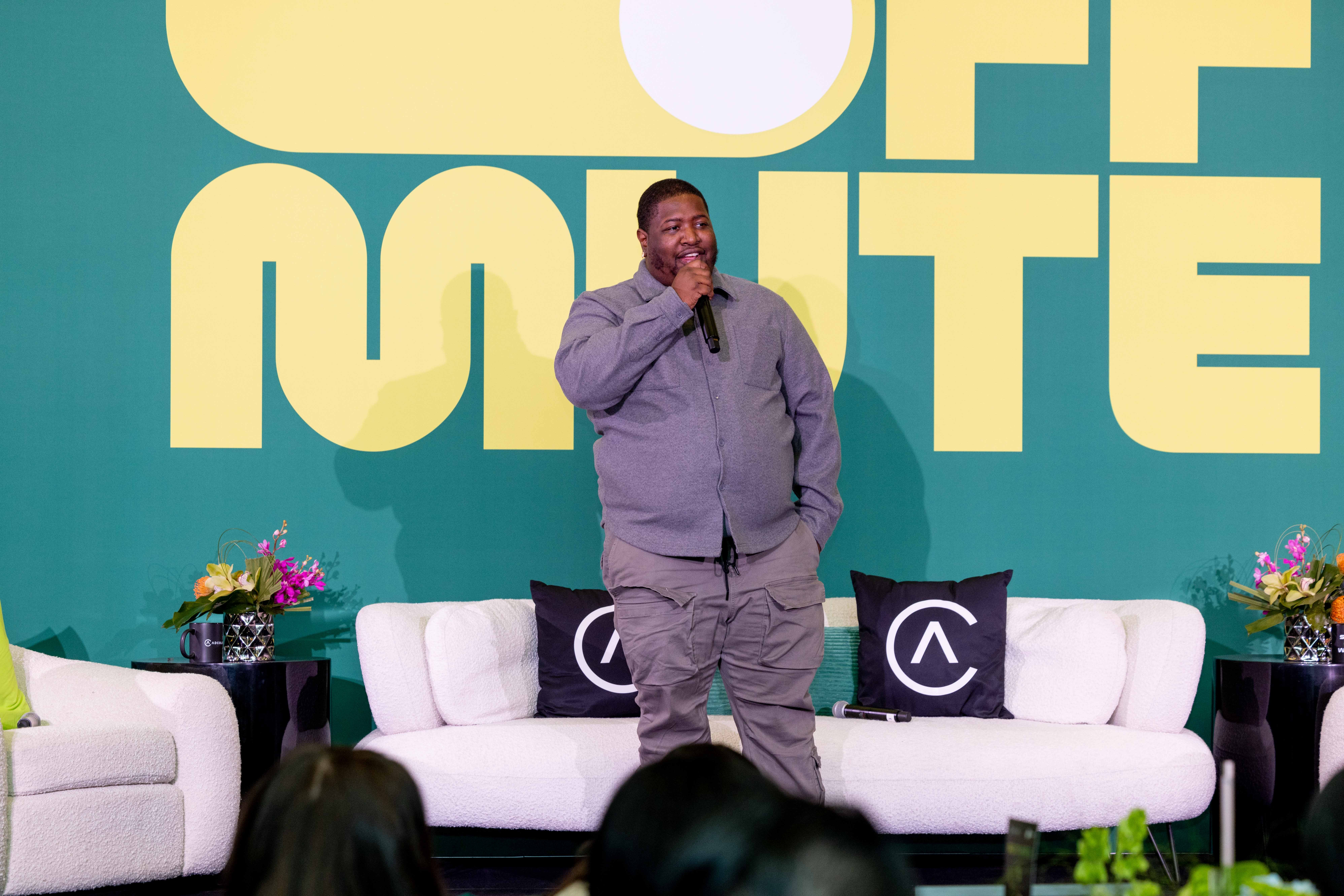 Man standing on stage with microphone, smiling, in front of large yellow text, flanked by white sofas and flowers.