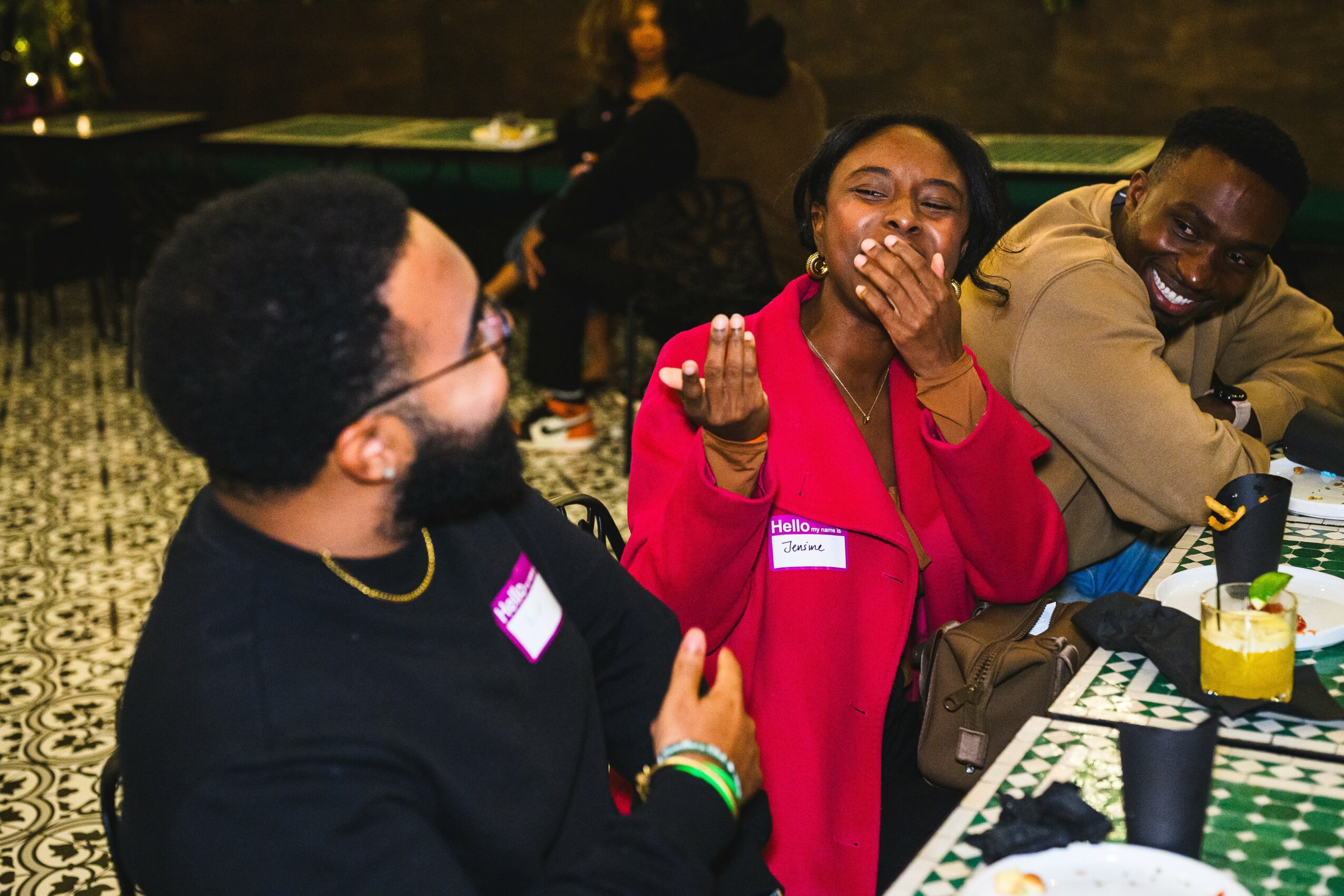 Three people sitting at a table, smiling and laughing, in a social setting with drinks and snacks.