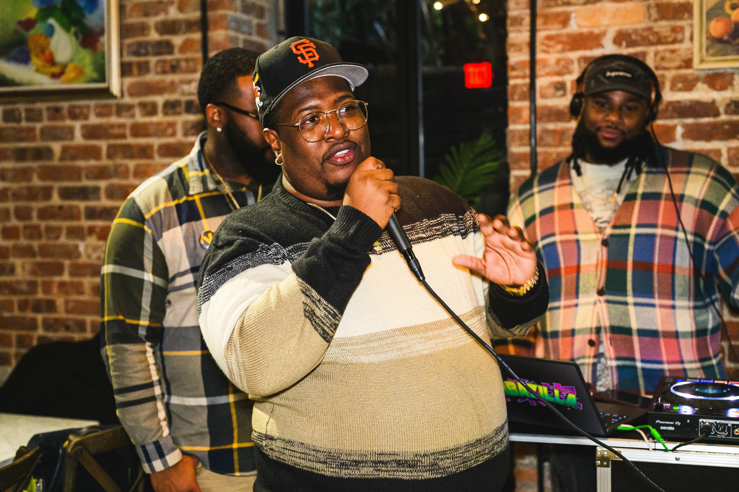 Three men in casual clothing and glasses, one speaking into a microphone, in a brick-walled room with a DJ setup.