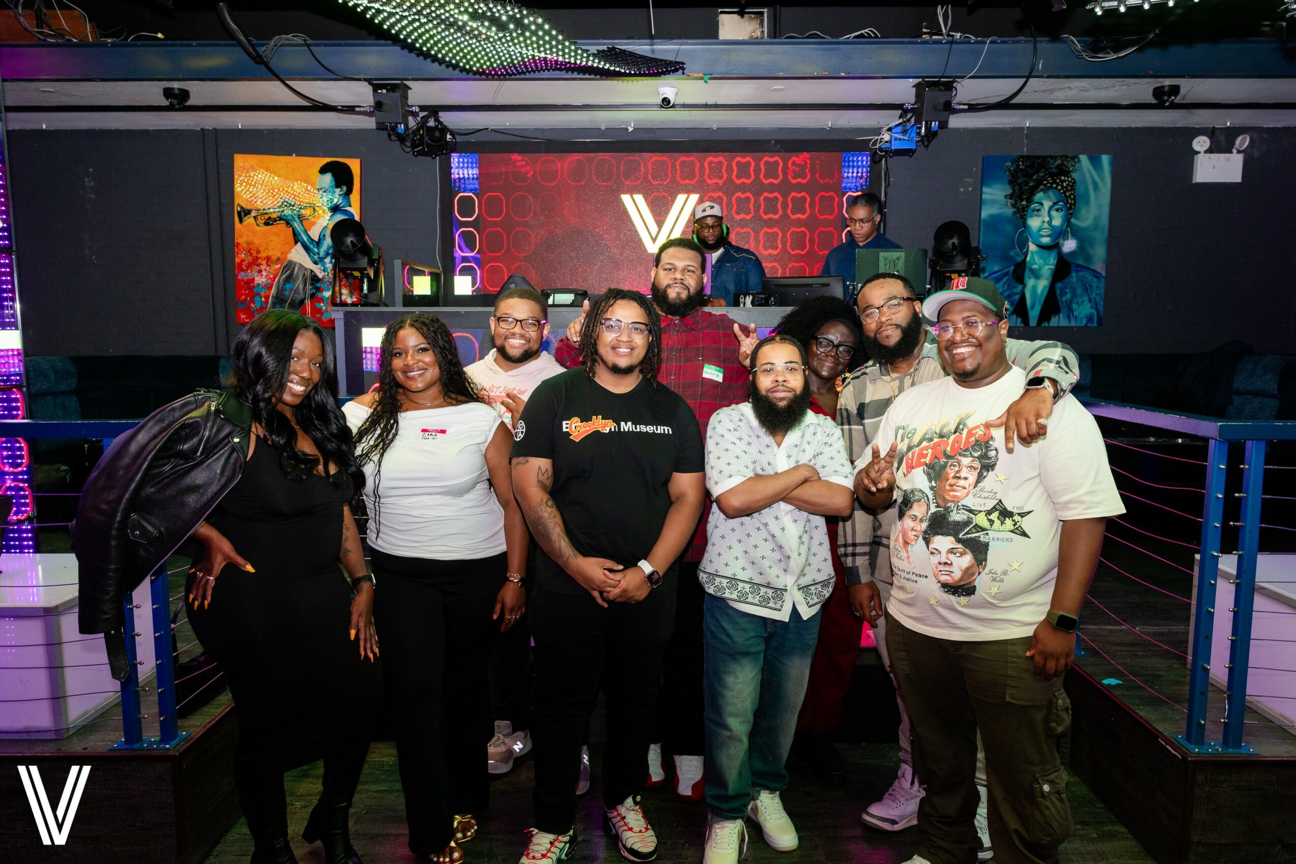 Group of ten people posing in a nightclub with colorful artwork and a DJ booth in the background.