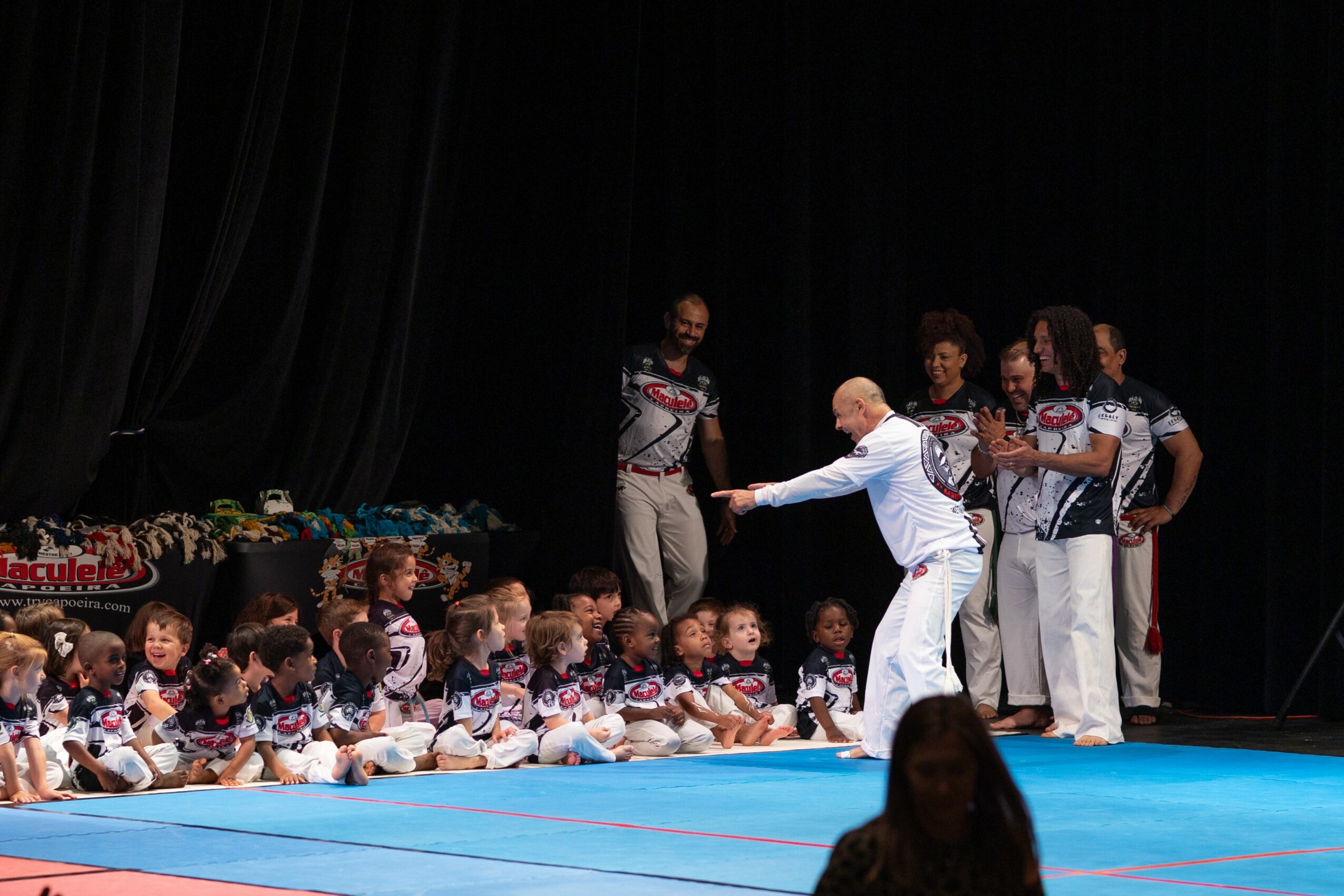 Group of adults and children on stage, children sitting on the floor, adults standing behind, stage with black curtains, blue mat.