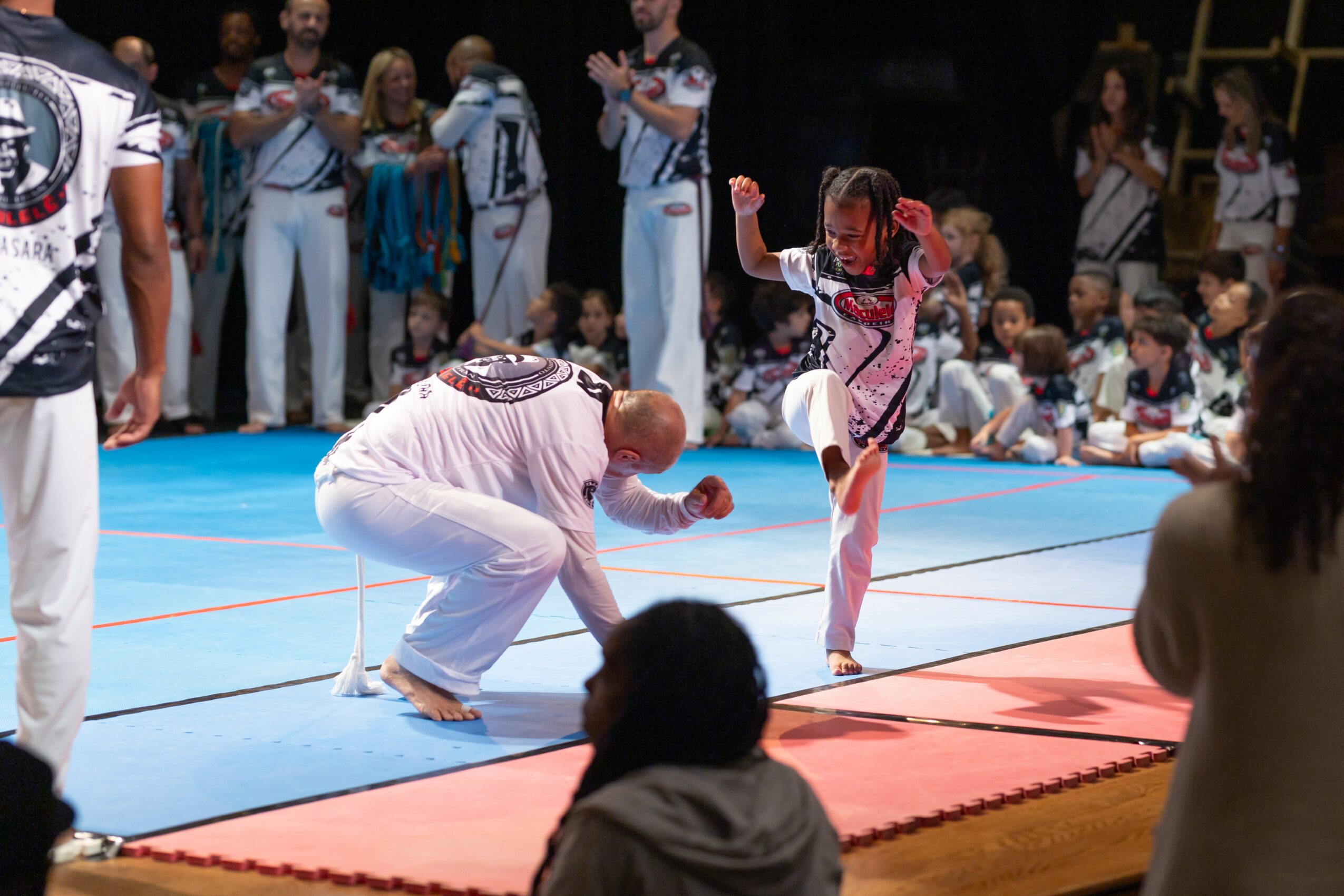 Martial arts demonstration with a person in a pink uniform performing a kick, surrounded by an audience and other participants.