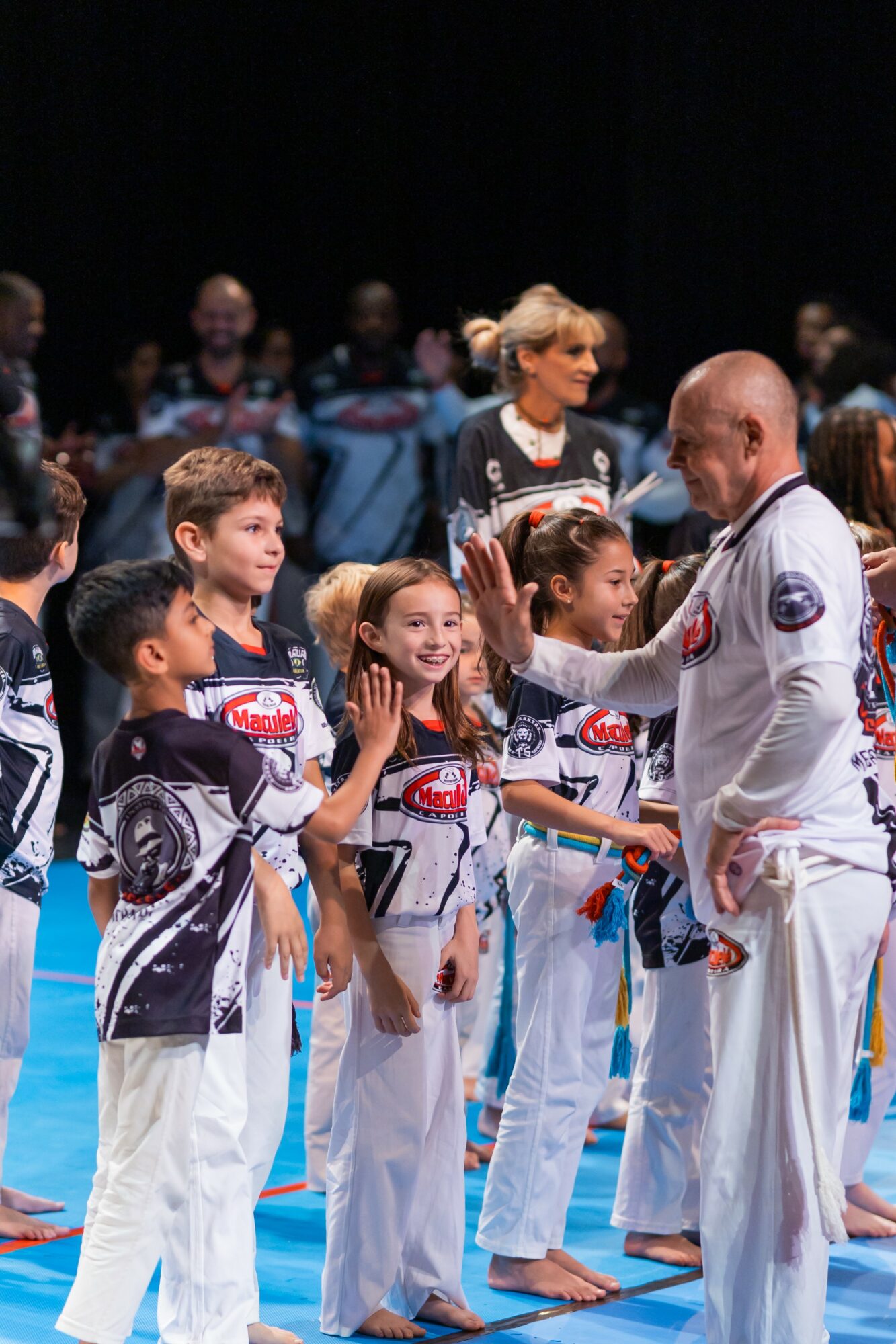 Children in martial arts uniforms receiving awards from an instructor, with a group of people in the background.