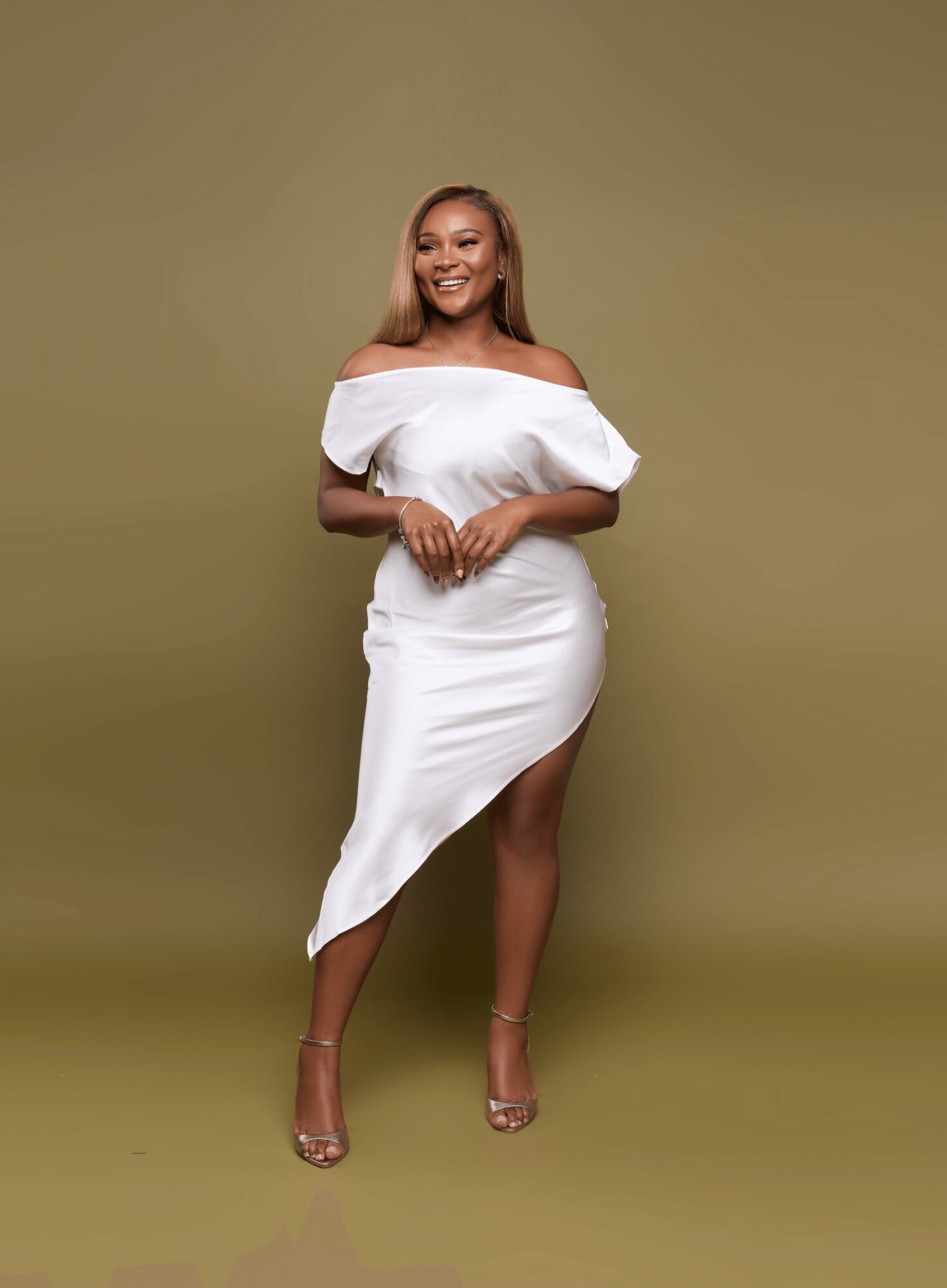 Smiling woman in white off-shoulder dress with slit, standing against olive green background.