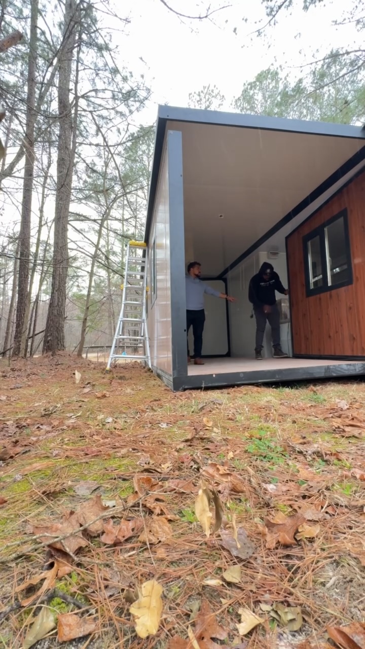 Two people standing inside a modern structure with a wooden wall, outdoors with trees in the background.