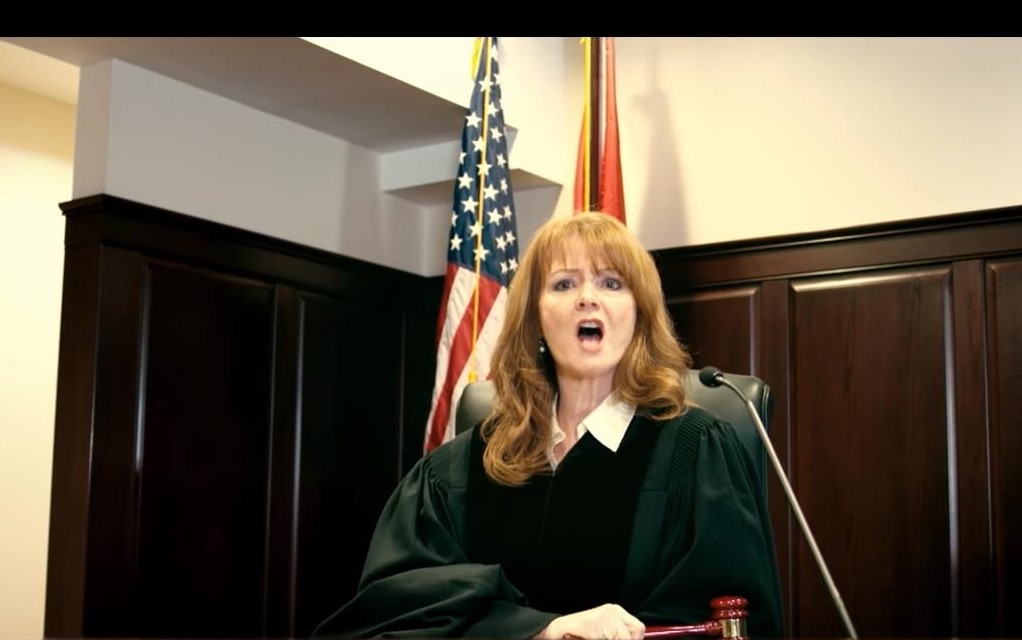 Female judge in black robe speaking in courtroom with American flag behind her.