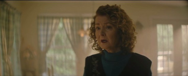 Woman with curly hair wearing dark clothing in a room with windows and curtains.