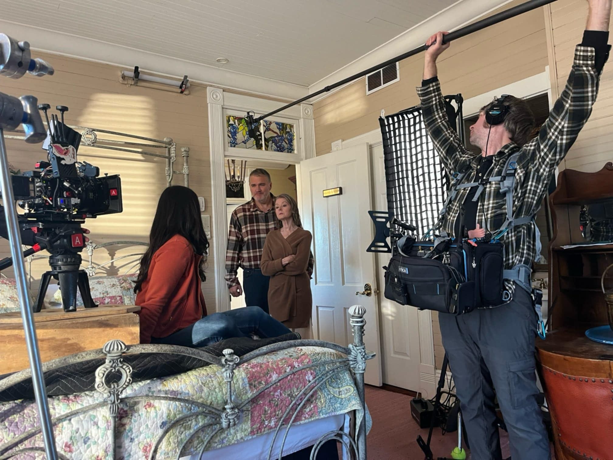 Filming crew records a scene with three actors in a room, with a person holding a boom microphone nearby.