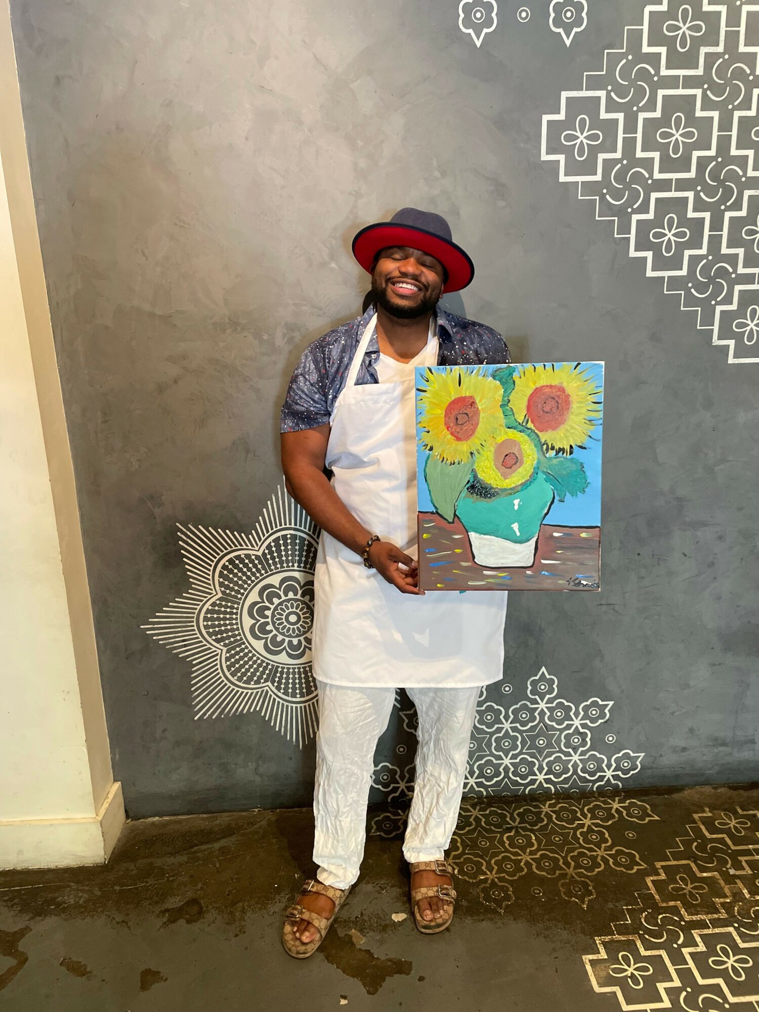 Person wearing a red hat and white outfit holding a colorful sunflower painting, standing against a gray wall.