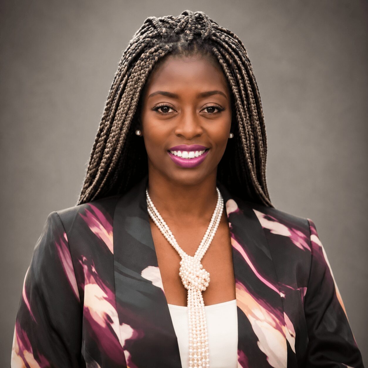 Portrait of a woman with long braided hair, wearing a floral blazer and pearl necklace, smiling against a plain background.