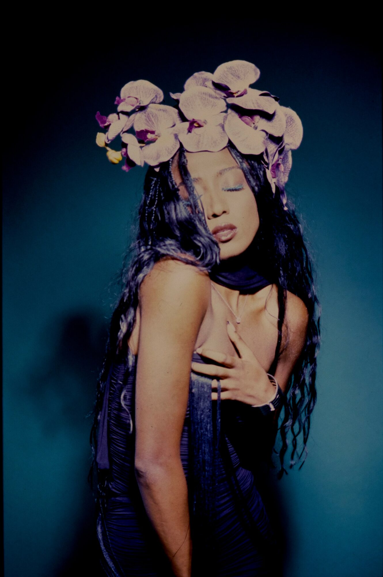 Woman with long dark hair wearing a large flower headpiece, eyes closed, hand on chest, dark background.