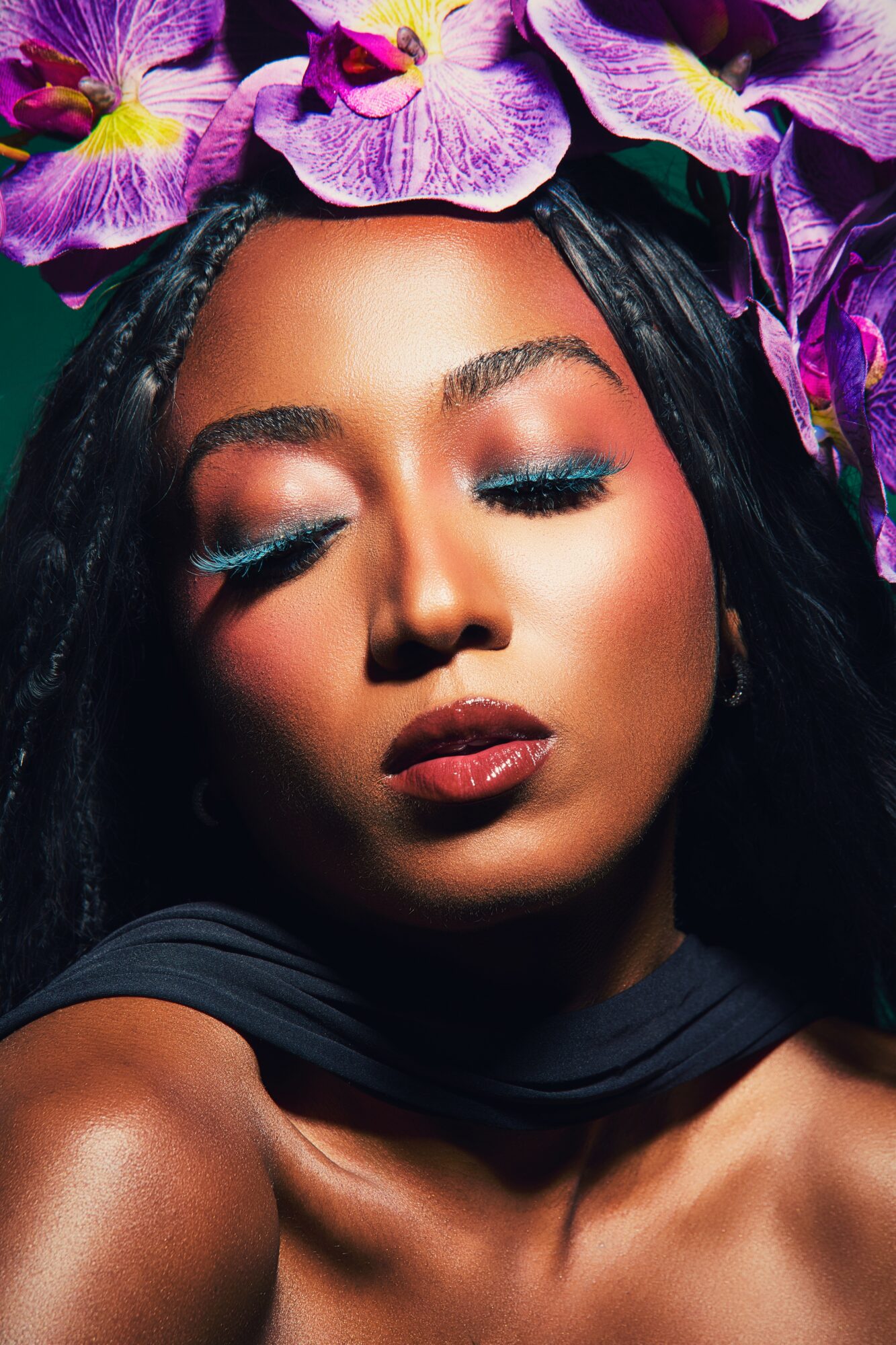 Close-up of a woman with closed eyes, vibrant makeup, and purple flowers in her hair.