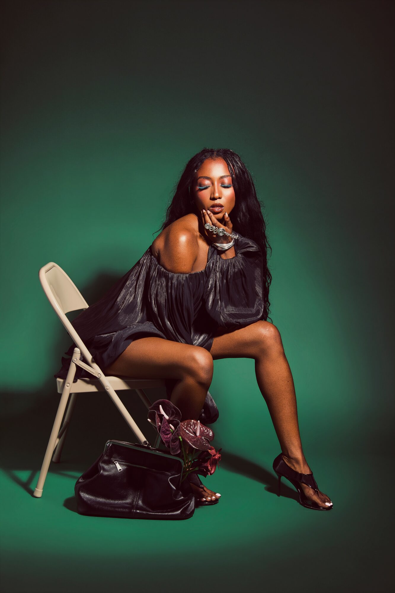 Woman sitting on a chair with a green background, wearing a black off-shoulder dress, with a bag and shoes nearby.