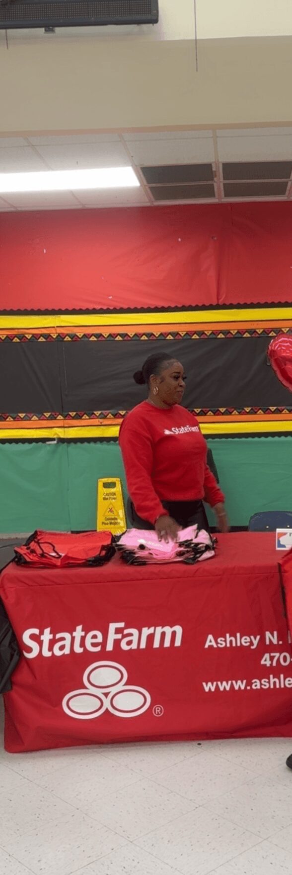 Person standing behind a table with a red tablecloth and State Farm banner, in an indoor setting.
