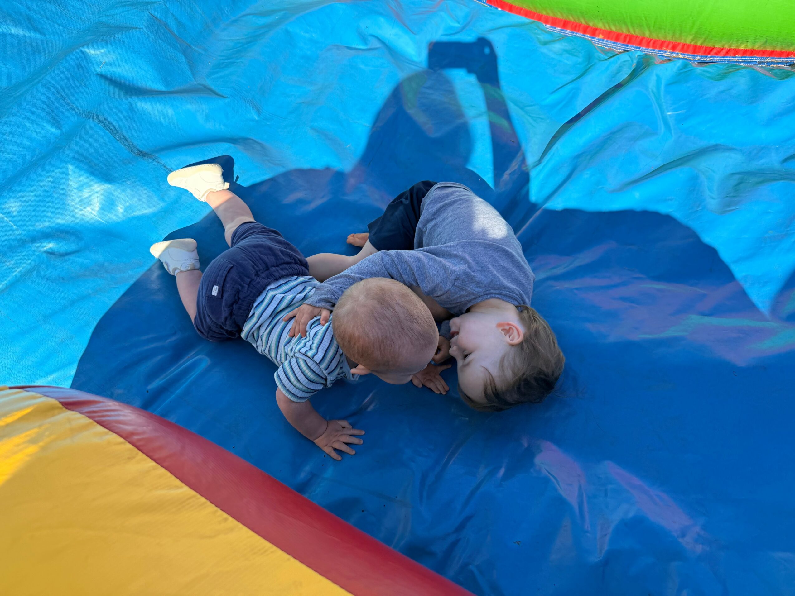 Two children lying on a colorful inflatable bounce house, playing and cuddling, with shadows cast on the surface.