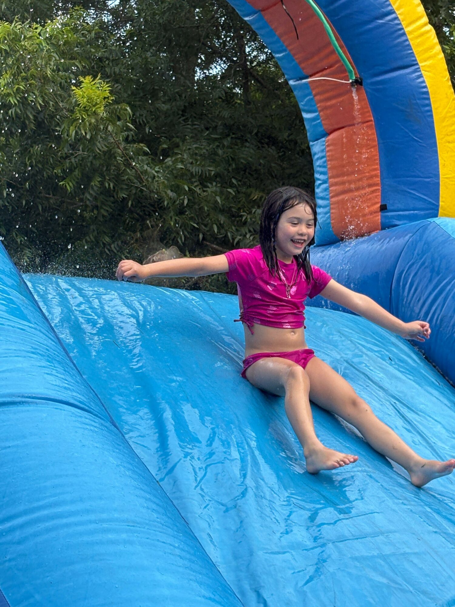 Girl in pink clothes lying on an inflatable water slide outdoors, with trees in the background.